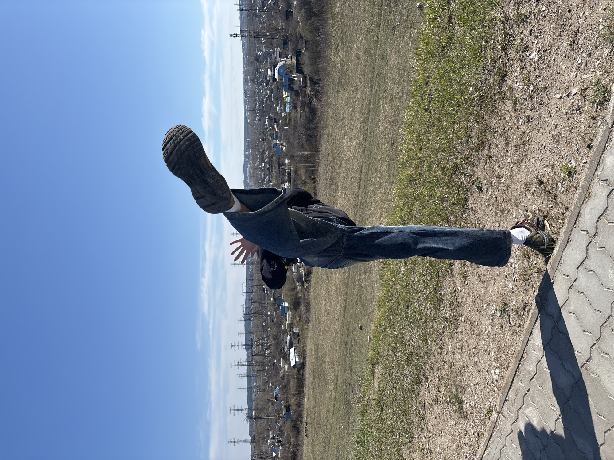 A person is positioned horizontally, suspended in the air. One leg, wearing a brown shoe, extends upward toward a clear blue sky with faint white clouds. The other leg, with a sneaker and visible white sock, is positioned toward a paved sidewalk and grassy ground. The person wears blue jeans, a dark jacket, and a black cap. Their arm is extended outward. In the background, there's a view of the city of Chaikovsky, Russia, characterized by residential buildings, power lines, and sparse vegetation, with a predominantly clear sky.