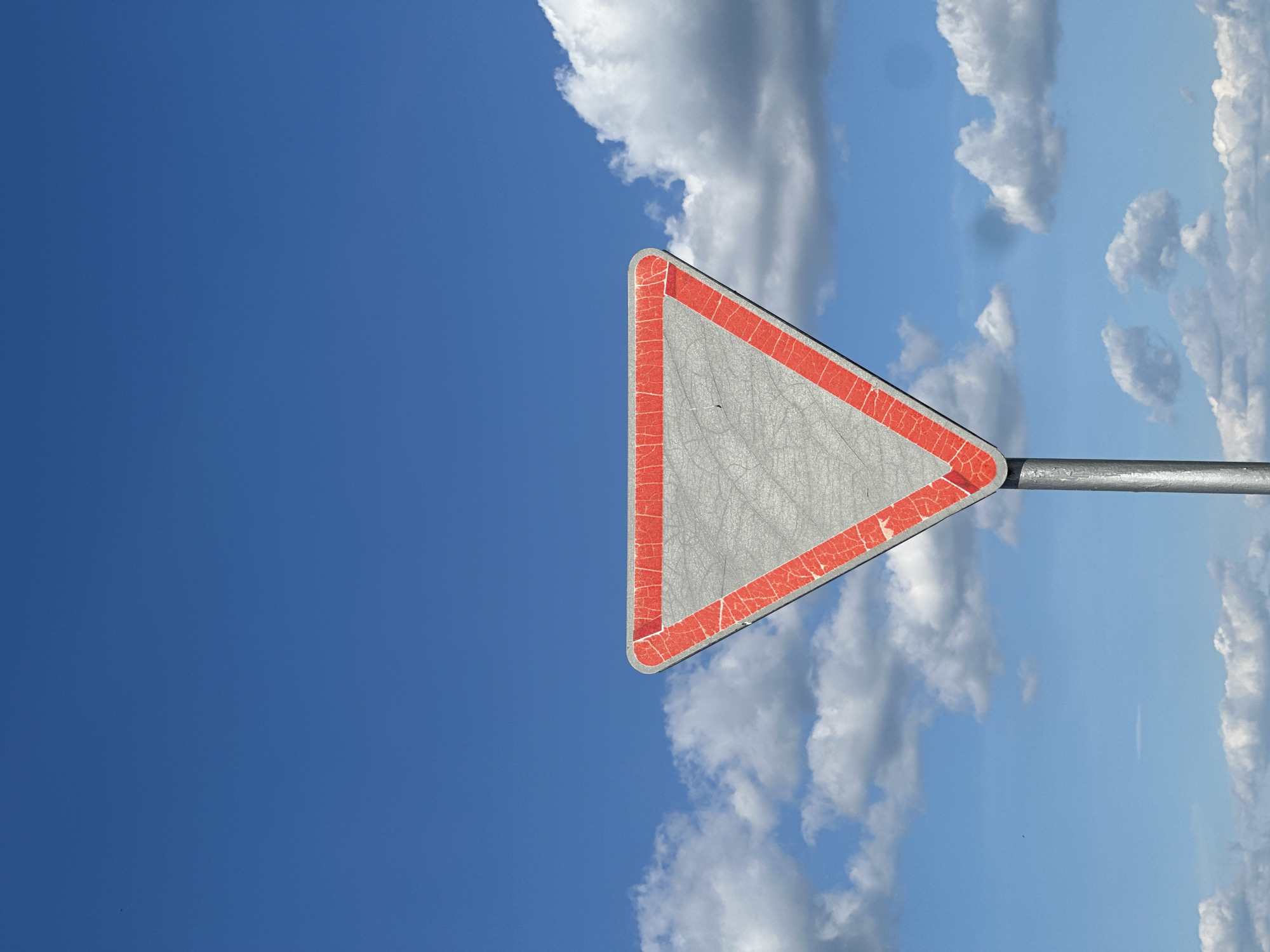 The image depicts a triangular traffic sign with a red border against a bright blue sky filled with fluffy white clouds. The sign is mounted on a metal pole, which extends horizontally across the lower right portion of the frame. The surface of the sign is a weathered grey and shows signs of cracking and wear along the red border. The sky is clear and vast, with sunlight illuminating the clouds and the sign. The orientation of the image is vertical. There is no visible text on the sign itself. The setting appears to be an outdoor location, likely a roadside in Belarus, given the provided location context. The overall mood is serene and expansive, with the vastness of the sky dominating the scene.