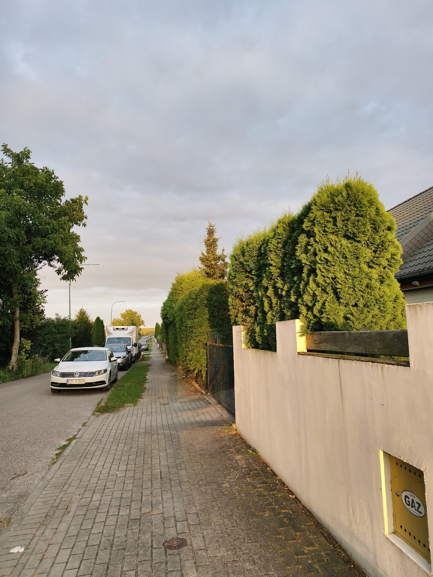 The image depicts a street scene in Tczew, Poland, during what appears to be late afternoon or early evening, given the lighting. On the left side of the image, a paved road is visible with a white Volkswagen sedan parked along its edge. The car's license plate reads "GTC 57028". To the right of the road is a grey brick sidewalk.
The right side of the image is dominated by a white stucco wall that runs along the property line. Mounted on this wall is a yellow gas meter box with "GAZ" inscribed on it, indicating a natural gas connection. A wooden fence is visible behind the stucco wall, partially obscured by a line of dense, green coniferous trees that form a hedge.
Further down the street, a line of vehicles, including a white delivery van and several cars, are parked along the side of the road, suggesting a residential area. Tall green trees and hedges line the sidewalk and the road, with a clear sky with scattered clouds overhead. The overall impression is a quiet, well-maintained residential street.