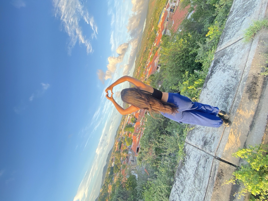 A young woman stands on a weathered stone wall, overlooking a cityscape. She's wearing a black top and blue pants, her long dark hair cascading down her back. With her arms raised, she forms a heart shape with her hands, framing the blue sky with scattered clouds. The city sprawls below, with buildings peeking through lush green trees. The scene is bathed in the warm light of the late afternoon or early morning sun, creating a serene and picturesque moment.