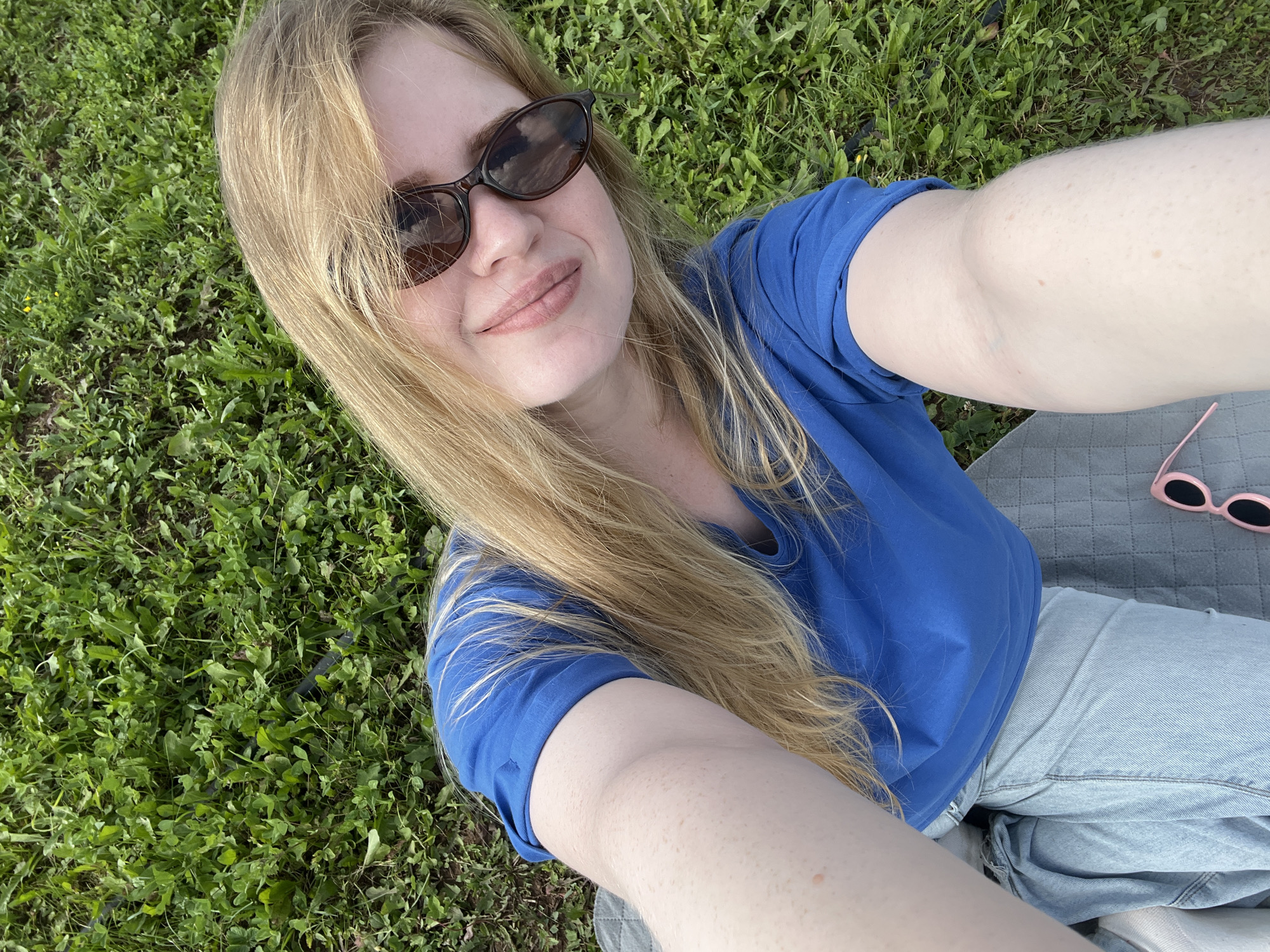 A young woman with long blonde hair and sunglasses is lying on a patch of grass in the city of Bolshoy Bukor, Russia. She is wearing a blue t-shirt and light-wash jeans. Her arms are extended to take a selfie, and she is smiling. The grass is green and lush, and there are a few small yellow flowers visible. A grey blanket is spread out beneath her, and a pair of pink sunglasses rests near her feet. The overall scene suggests a casual, outdoor setting, possibly a park or a backyard.
