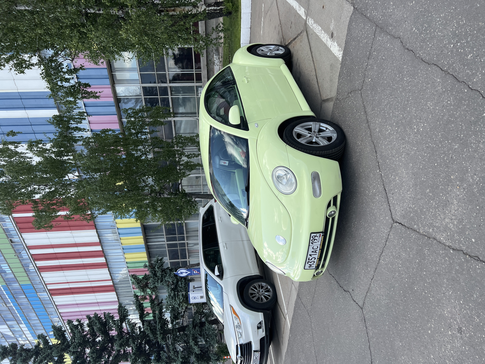 The image shows a light green Volkswagen Beetle parked on asphalt next to a white SUV. The Beetle has a Russian license plate reading "M351AC 199 RUS". Both cars are parked near a building with a distinctive facade of horizontal bands of color, including red, white, blue, yellow, green, and pink. The background includes trees. A sign is visible near the white SUV. This scene is likely located in Khimki, Russia.