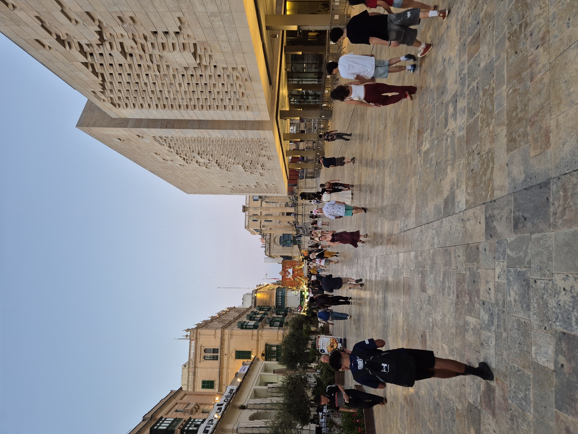 This image captures Republic Street in Valletta, Malta, during dusk. The street is paved with large, irregular stone tiles and is bustling with pedestrians walking in both directions. On the right side of the street, the modern building of the Parliament House, designed by Renzo Piano, dominates the scene with its distinctive honeycomb-like stone facade. The building features large glass windows on the lower level, suggesting a public space or entrance. On the left side, older, traditional Maltese buildings with yellow facades, wooden balconies, and green shutters line the street.
A diverse group of people are seen in the image, dressed in casual summer attire. Some individuals are walking alone, while others are in pairs or small groups. The lighting suggests the transition from daylight to evening, with a soft, ambient glow illuminating the street and casting subtle shadows. Towards the end of the street, a festive decoration with lights and banners is visible, indicating a possible event or celebration. The sky is a clear, pale blue, characteristic of twilight.