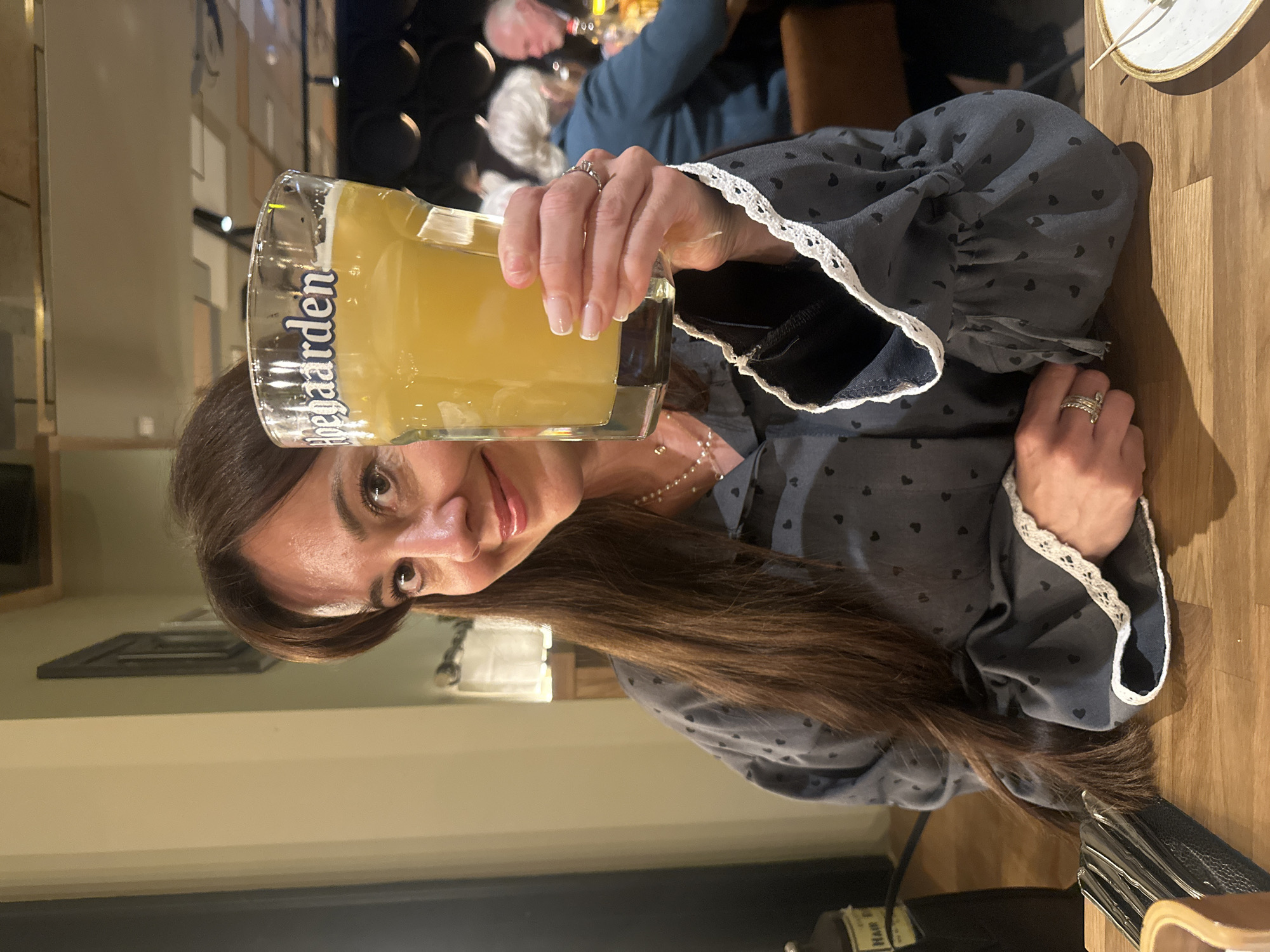 A woman sits at a wooden table holding a full glass of beer with the word "Hoegaarden" printed on it in Riga, Latvia. The woman has long brown hair and is wearing a patterned grey shirt with white lace on the sleeves. She is smiling at the camera. In the background, other people are seated at tables in a restaurant. A plate is also visible on the wooden table.