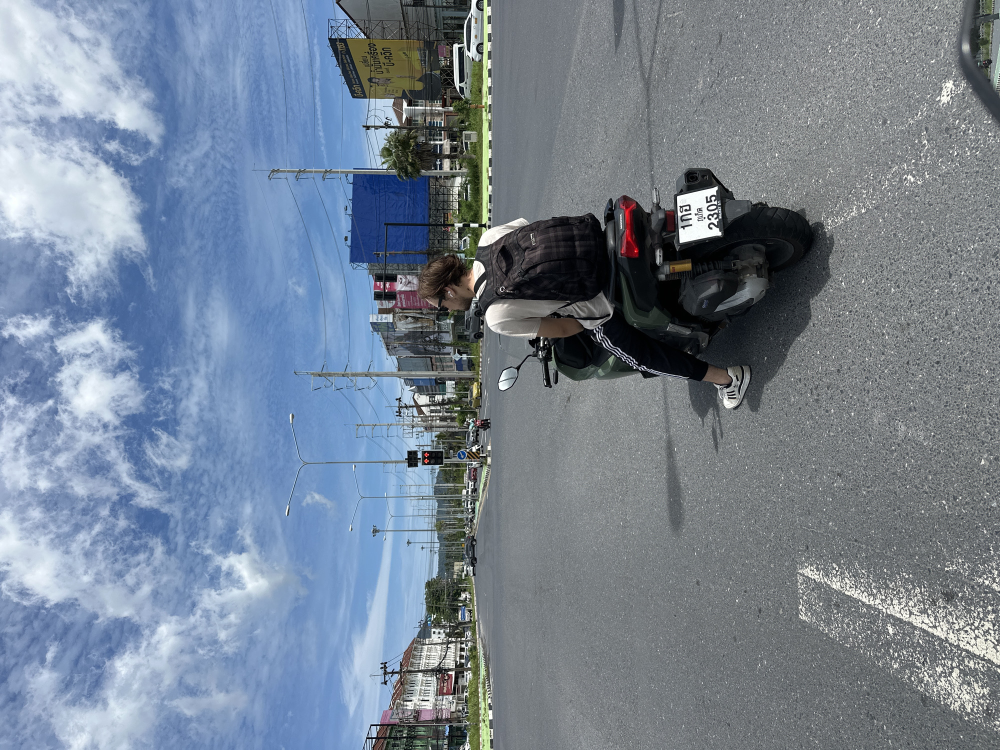 In a city scene, a person with light brown hair and wearing sunglasses is seated on a dark green motorcycle. They have a dark plaid backpack on their back and are dressed in a light-colored t-shirt and black athletic pants with white stripes. The motorcycle has a license plate with Thai characters and the numbers "2305". The person is on a paved road, appearing to be at an intersection, with traffic lights visible in the background. The environment includes buildings, utility poles with wires, streetlights, billboards, and other vehicles. The sky is blue with scattered white clouds.