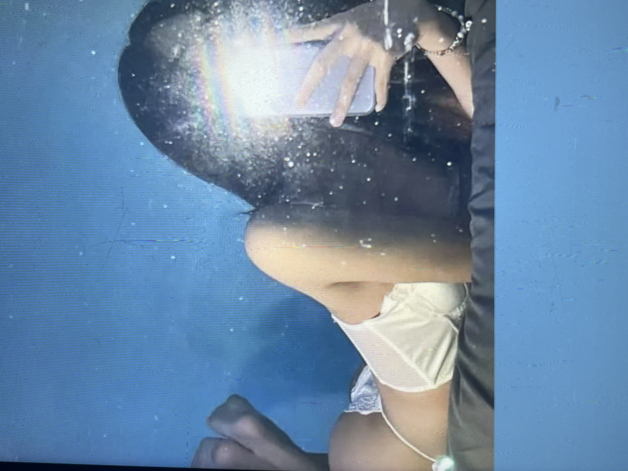 This is a low-quality, pixelated image of a person lying on their stomach, facing away from the camera, on a dark gray surface. They are wearing white lingerie and have long dark hair. Their left foot is bent upwards. In the background, the wall is a bright blue. The person is holding a phone in their right hand, taking a selfie. The phone screen is bright and reflects a rainbow-like glare. There are small white specks scattered across the image, possibly dust or a screen artifact. The image is unclear and lacks sufficient detail to determine a specific location within Bishkek, Kyrgyzstan, or to provide further forensic information.