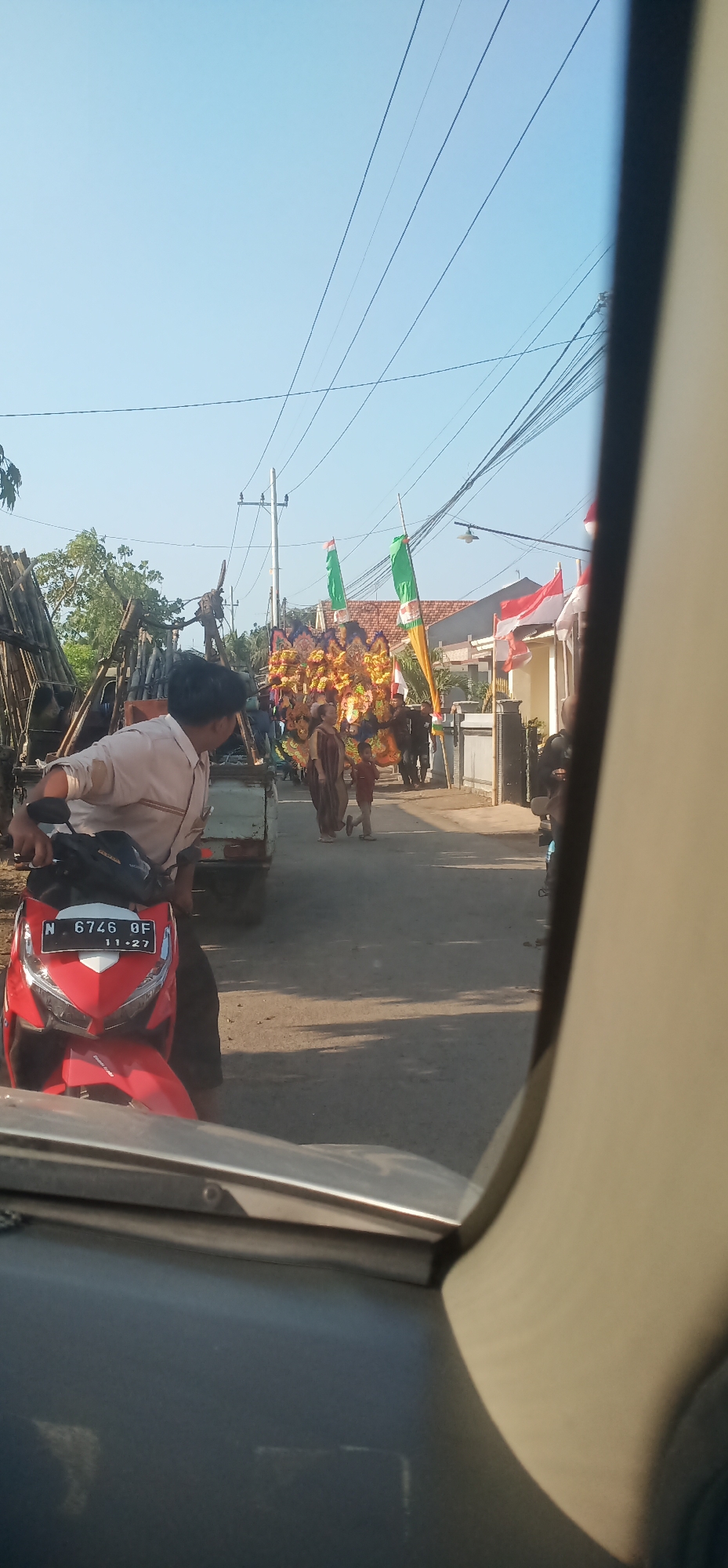 A parade is taking place on a street in Randuputih, Indonesia. The procession includes a large, decorated float on a truck, adorned with gold and colorful fabric. Several people are walking alongside the float, some dressed in traditional clothing. Two green flags and one red and white Indonesian flag are visible, attached to poles and waving in the breeze. On the left side of the street, a red motorcycle with license plate "N 6746 QF" is parked. A person wearing a light-colored shirt is seated on the motorcycle, looking towards the parade. The street is paved, and there are houses and trees lining the sides. Overhead, power lines crisscross the clear blue sky. The scene appears to be captured from inside a vehicle, as indicated by the reflection and framing in the foreground.