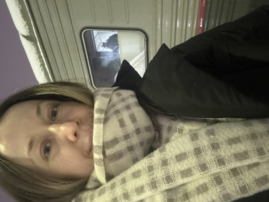 A person with light brown hair and a patterned scarf is facing the camera. A dark-colored jacket is draped to the right. Behind them is the side of a white, possibly metallic structure with narrow vertical ridges. A small window is visible, showing a dark figure inside the structure, giving the impression they might be on a vehicle or within a building. The sky in the upper left corner has a pale, lavender hue.