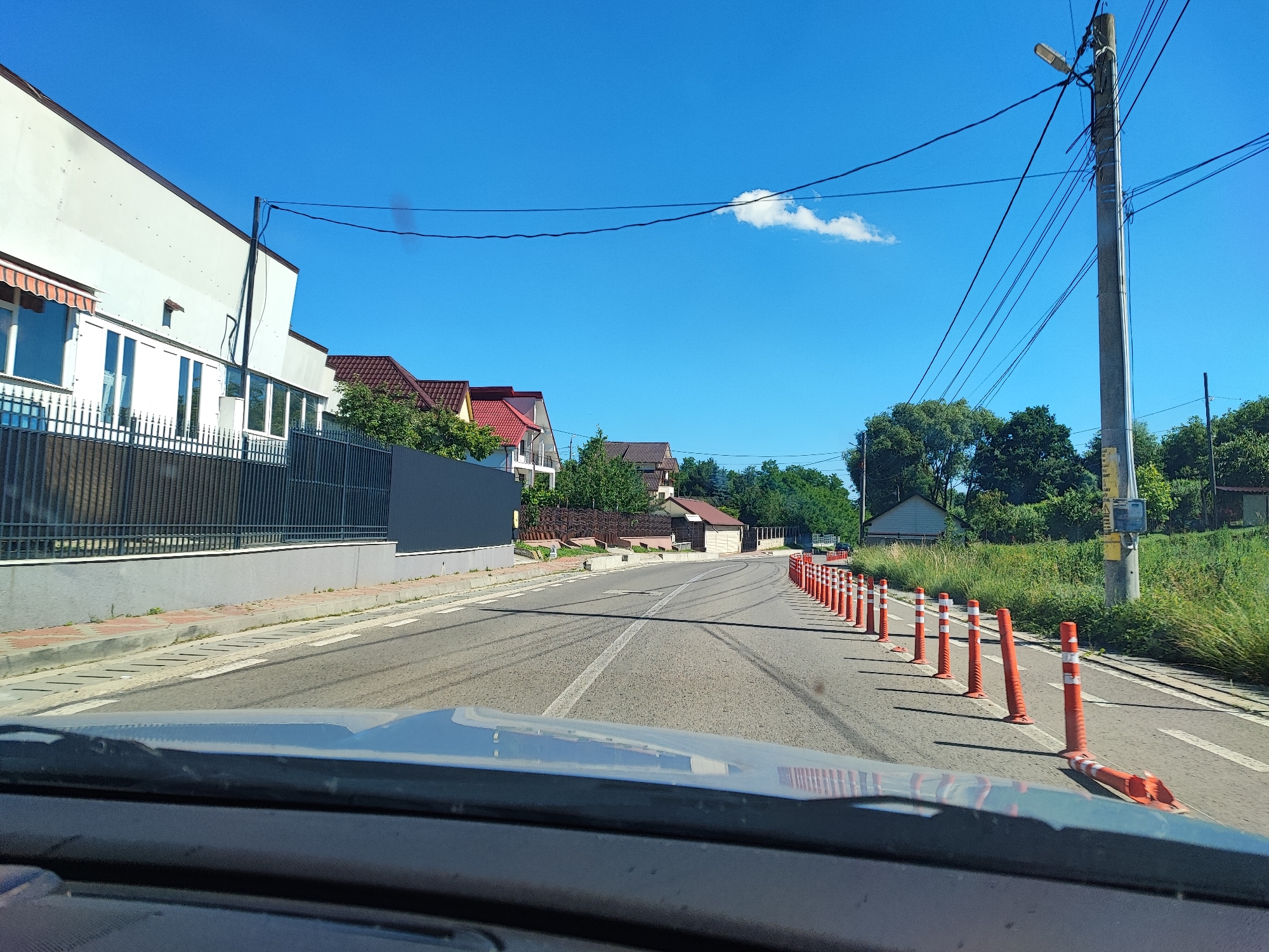 The image depicts a road in Târgu Neamț, Romania, on a clear, sunny day. The perspective is from inside a vehicle, looking forward. The road is paved and has a white line marking the center. On the right side of the road, a long row of orange traffic cones, some standing upright and others leaning or lying down, are placed along the edge. Beyond the cones, there is a grassy area with tall weeds and trees. On the left side of the road, there is a building with a white facade, a dark fence, and a striped awning. Further down the road, several houses with red-tiled roofs and trees are visible. A utility pole with multiple wires stretches across the scene, with a street lamp attached to it. The sky is bright blue with a single, small white cloud.