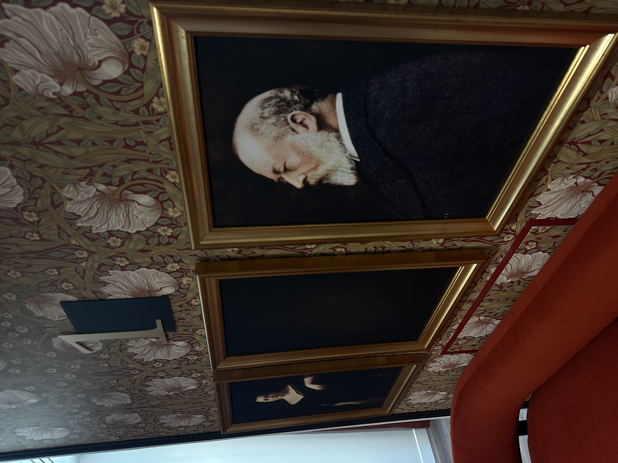 The image captures a scene in a Parisian interior. Three framed portraits hang on a wall adorned with a William Morris-style floral wallpaper. The top portrait depicts an elderly man with a beard, his head bowed. The middle portrait is dark and less distinguishable. The bottom portrait shows a woman in a dark dress. A black, angular object is attached to the wall. The corner of an orange upholstered object, likely furniture, is visible on the right. A window with sheer curtains is at the bottom.