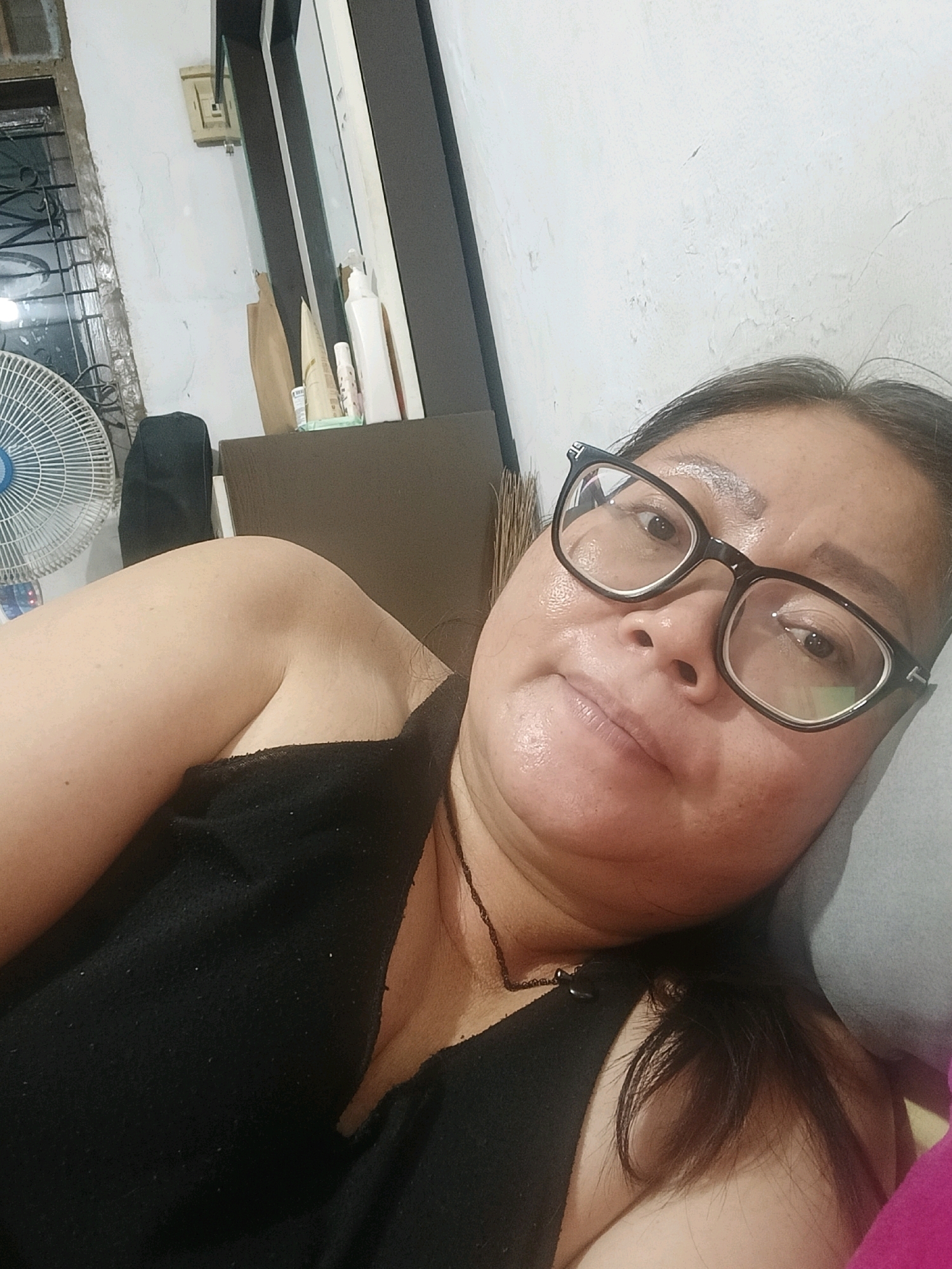 A middle-aged woman with dark hair and glasses is lying down, taking a selfie. She is wearing a black top. She is in a room with a white, textured wall. To her left, there is a white oscillating fan and a window with metal bars. On a shelf behind her, there are several bottles and containers. The lighting suggests an indoor environment. The location is Jatimulya, Indonesia.