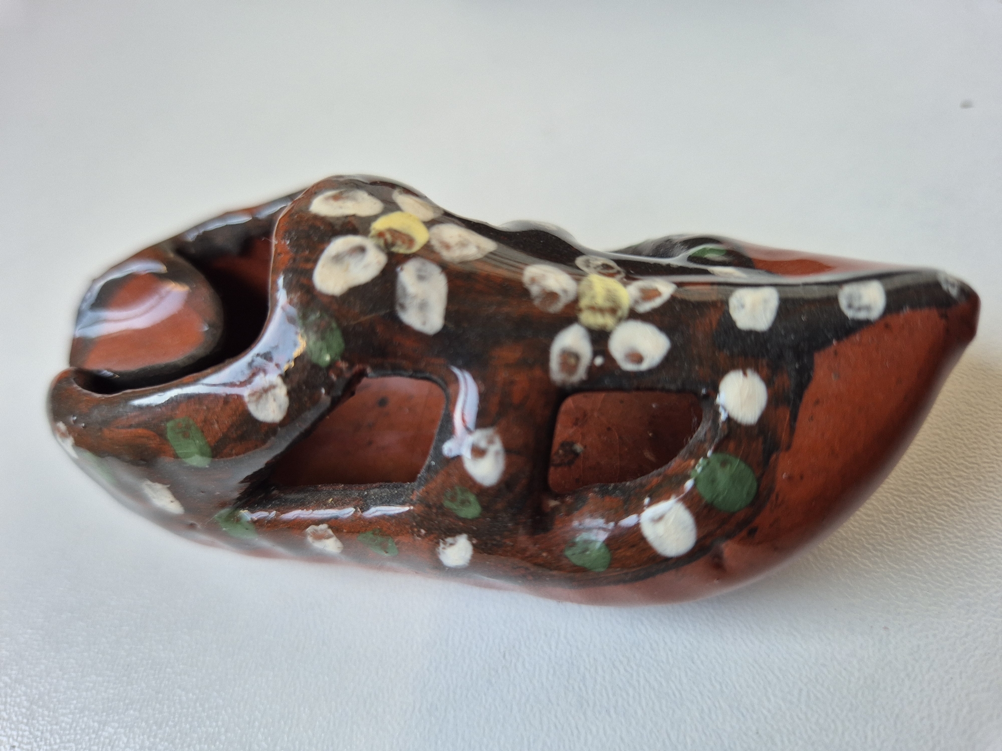 A small, brown, clay-like object with floral decorations is centered in the frame. The object has holes cut through its sides and appears to be glazed. The decorations include white and yellow floral patterns and green leaf-like shapes. The background is a plain, white surface.