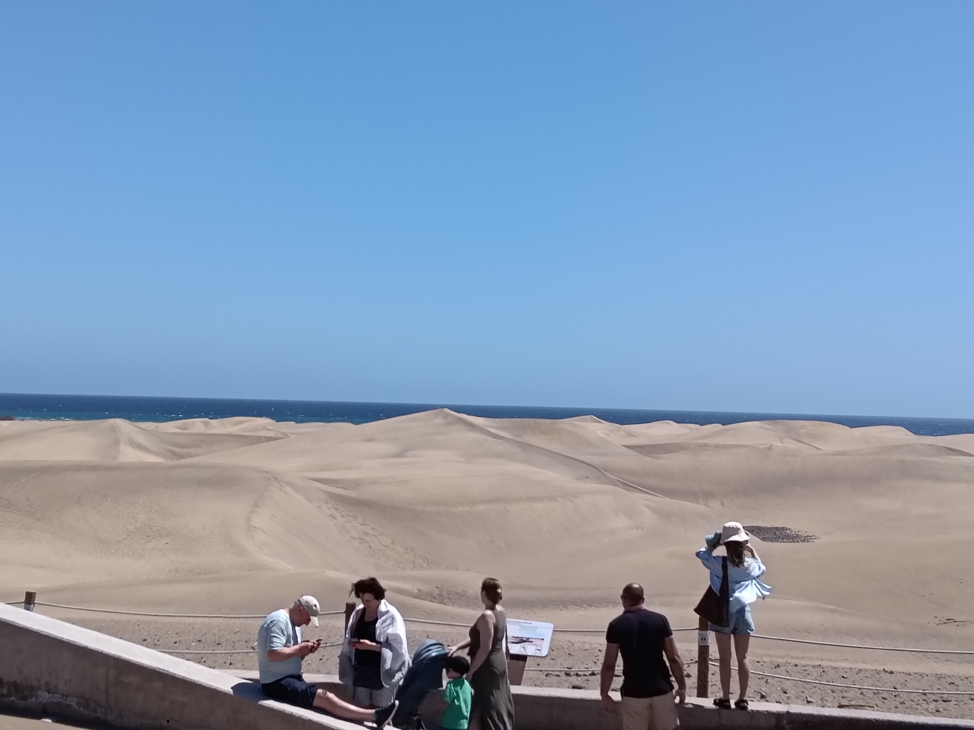 The image captures a scene at the Dunas de Maspalomas in San Bartolomé de Tirajana, Spain. The environment is dominated by large, undulating sand dunes that stretch towards the ocean horizon, separated by a clear blue sky.
Several people are present. From left to right, a man sits on a concrete barrier looking at a mobile phone, while a woman beside him is looking at her own phone; they are beside a baby stroller. Next to them, a woman in a long dress stands, watching a small child in a green shirt. To the right, a man is standing and facing the desert with his back turned to the camera. Finally, a woman in a hat and a light blue shirt stands at the edge, appearing to take photos or observe the scenery.
A concrete barrier with a rope fence divides the foreground from the sand dunes.