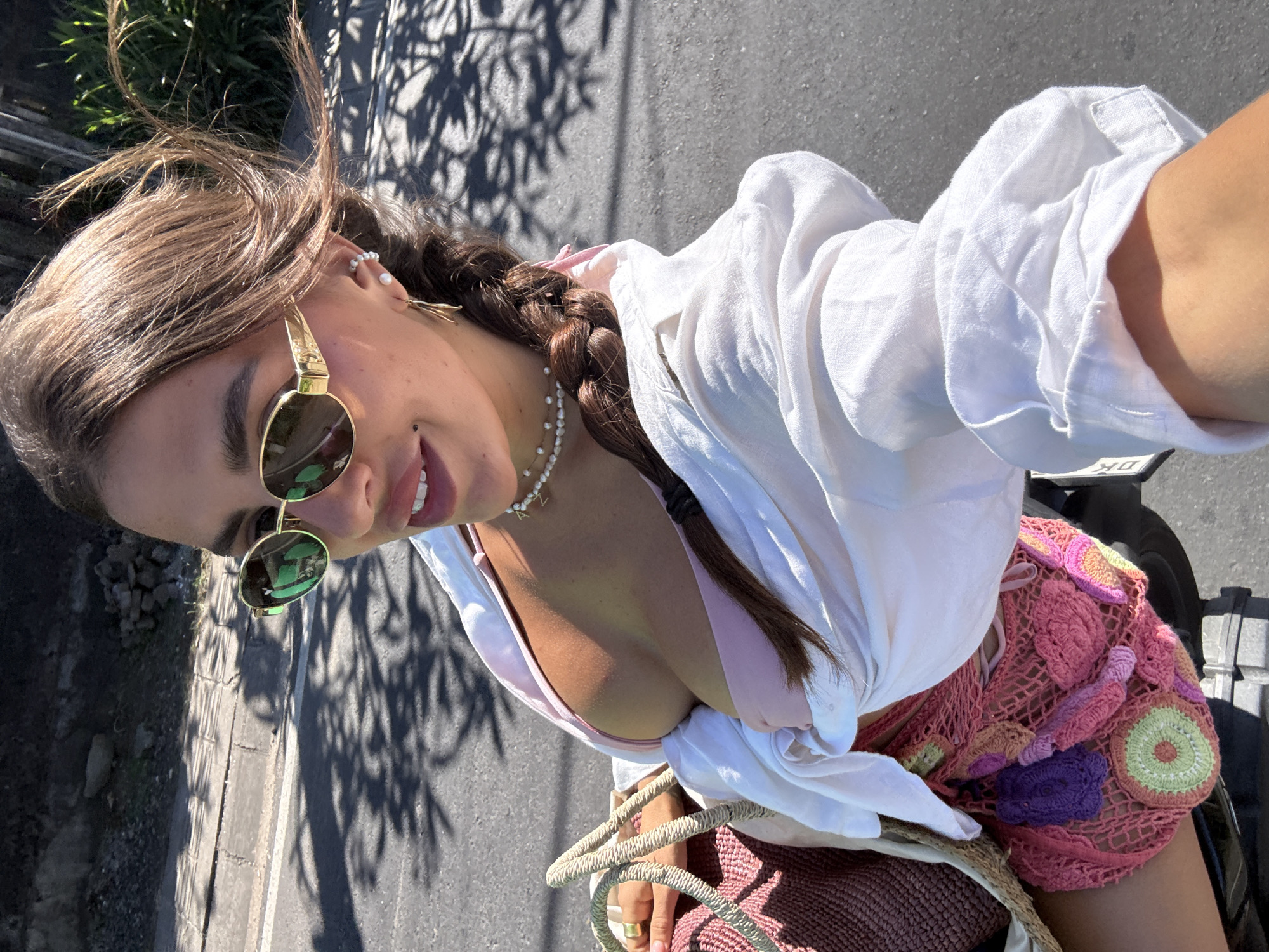 A woman is depicted from an elevated angle, presumably a selfie taken while on a scooter, given the scooter elements visible in the background and the angle of the shot. She wears round sunglasses with gold frames, a white shirt, a crocheted pink top, and a pearl necklace with a letter pendant. Her hair is styled in a braid. The background shows a paved road with shadows cast from trees, likely indicating a sunny day. Based on the visual cues, it is difficult to determine the exact location within Kuta Selatan without more specific landmarks or identifying features. The overall scene suggests a casual, summery setting.