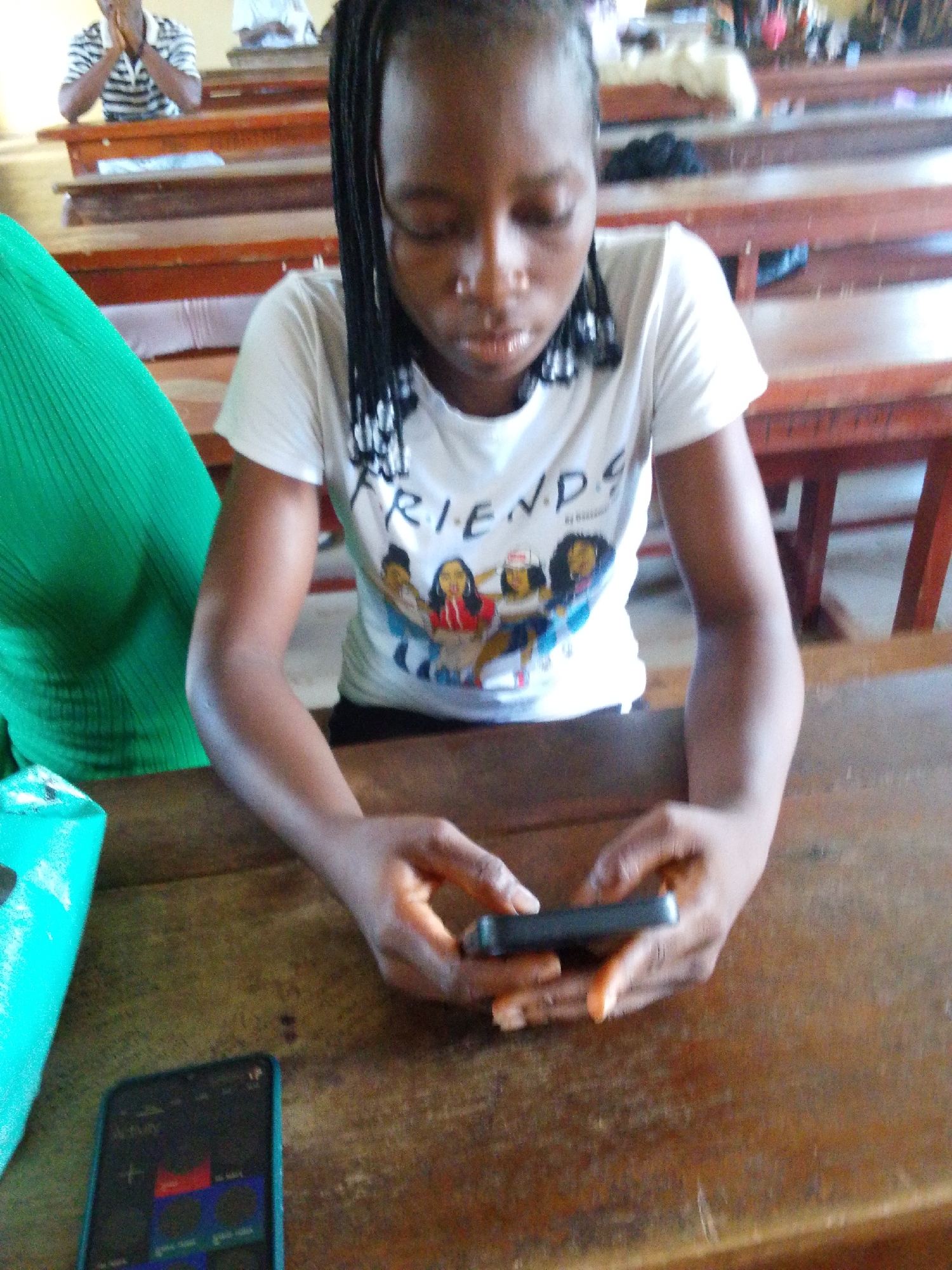 A young Black woman is seated at a wooden desk in an educational setting, possibly a classroom or lecture hall in the city of Unknown, Unknown. She is wearing a white t-shirt with a graphic of female friends and the word "FRIENDS" printed on it. Her hair is styled in braids. She is looking down and holding a dark-colored smartphone in her hands, with her fingers actively engaged with the device. Another smartphone is lying on the desk to her left. A woman in a green top is partially visible to her left. In the background, several rows of desks are visible, and two individuals are seated at one of the desks. The lighting appears to be natural, suggesting the presence of windows or an open space. The general environment suggests an academic or learning atmosphere.