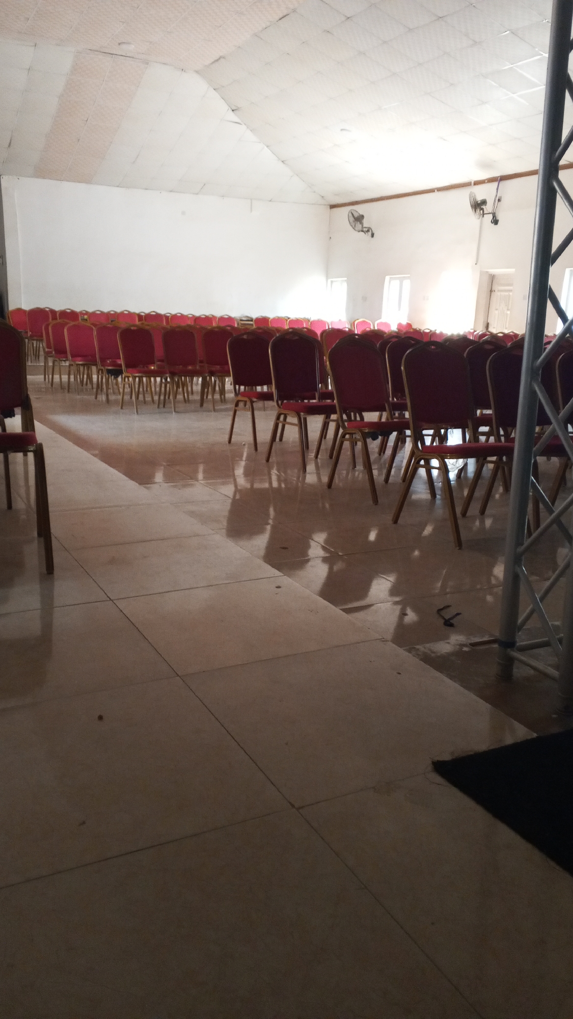 This media file depicts the interior of a large room with a high, sloped ceiling. The room is set up with rows of empty chairs, suggesting it's prepared for an event or gathering. The chairs are a deep red color with gold-colored metal legs and frames. They are arranged neatly in multiple rows, facing towards the front of the room, which is not fully visible.
The walls are painted white and are bare except for two oscillating fans mounted on the wall and some windows that admit natural light. The floor is tiled in a light, neutral color with a reflective sheen, showing shadows from the chairs and a metal support structure on the right side of the frame. The overall lighting suggests it might be daytime, with sunlight coming through the windows and reflecting off the floor.
There are no people visible in the image, and no specific activity is taking place, as the room is empty. The scene conveys a sense of anticipation or preparation for an event. The setting appears to be a hall, potentially a conference room, auditorium, or a gathering space within a community center or place of worship. There is no visible text in the image.