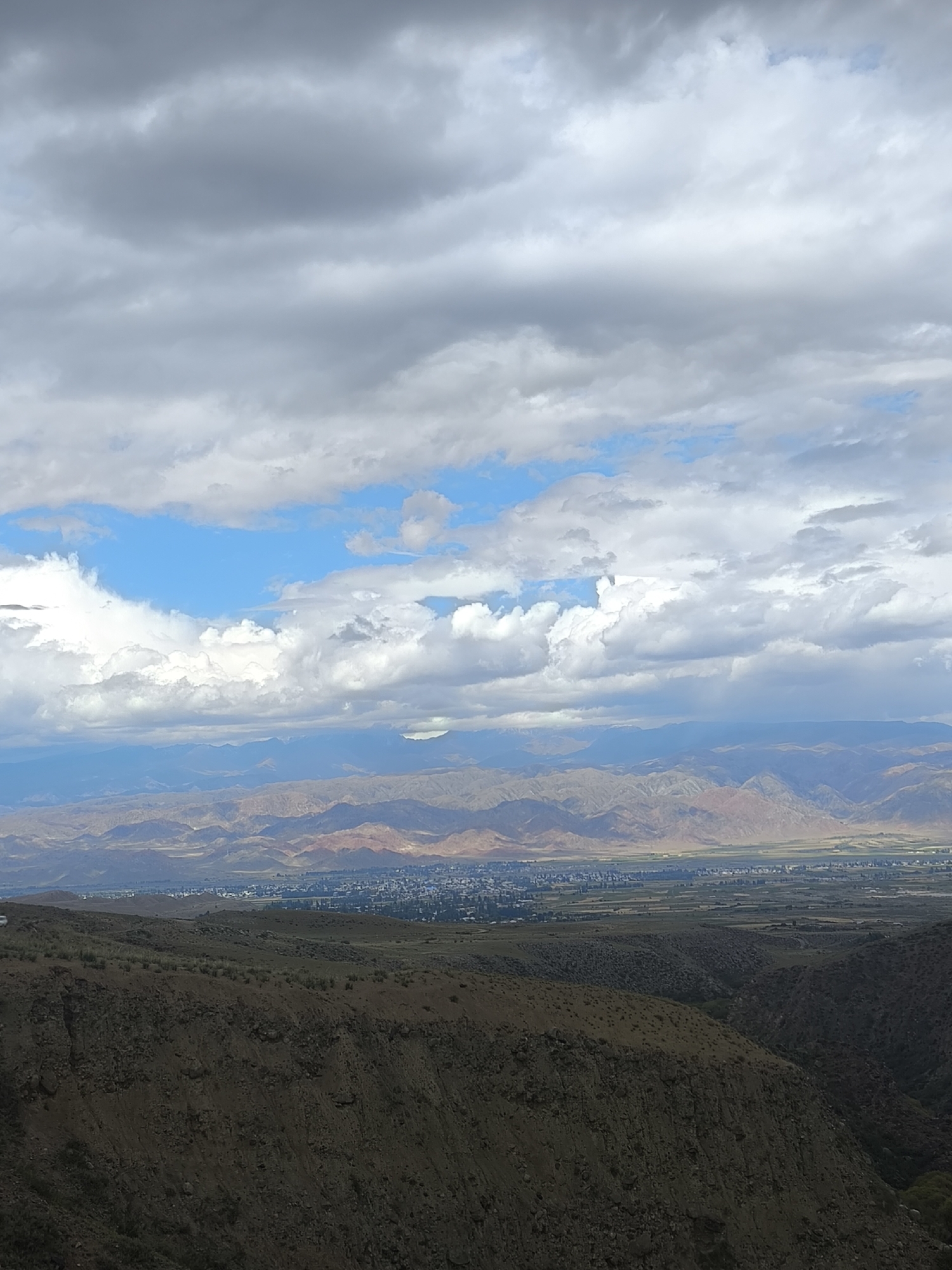 A wide, high-angle landscape shot captures a vast, mountainous terrain under a dramatic, cloud-filled sky. The foreground is dominated by a steep, rocky hillside covered in sparse vegetation. Beyond this, the middle ground reveals a valley with a town or city nestled within it, featuring buildings with blue roofs and green vegetation. Further in the distance, rolling hills and layers of mountains fade into the haze, with the highest peaks showing hints of snow. The sky is a mix of bright blue patches and heavy, gray clouds. The image does not contain any people or identifiable actions.