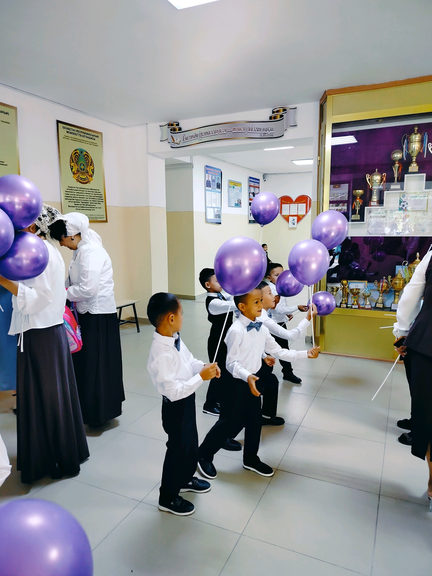 The image depicts an indoor scene, possibly a school hallway or event space, with a group of children holding purple balloons. The children are dressed in white shirts and dark pants, some wearing bow ties, suggesting a formal or celebratory occasion. They are standing and walking in a line, their faces turned slightly to the side. In the background, there are informational posters on the wall, one with the emblem of Kazakhstan. To the right, a display case filled with trophies and awards is visible. The floor is tiled, and the lighting appears to be artificial. The location is identified as Көлкент, Kazakhstan, indicating a setting within that city.