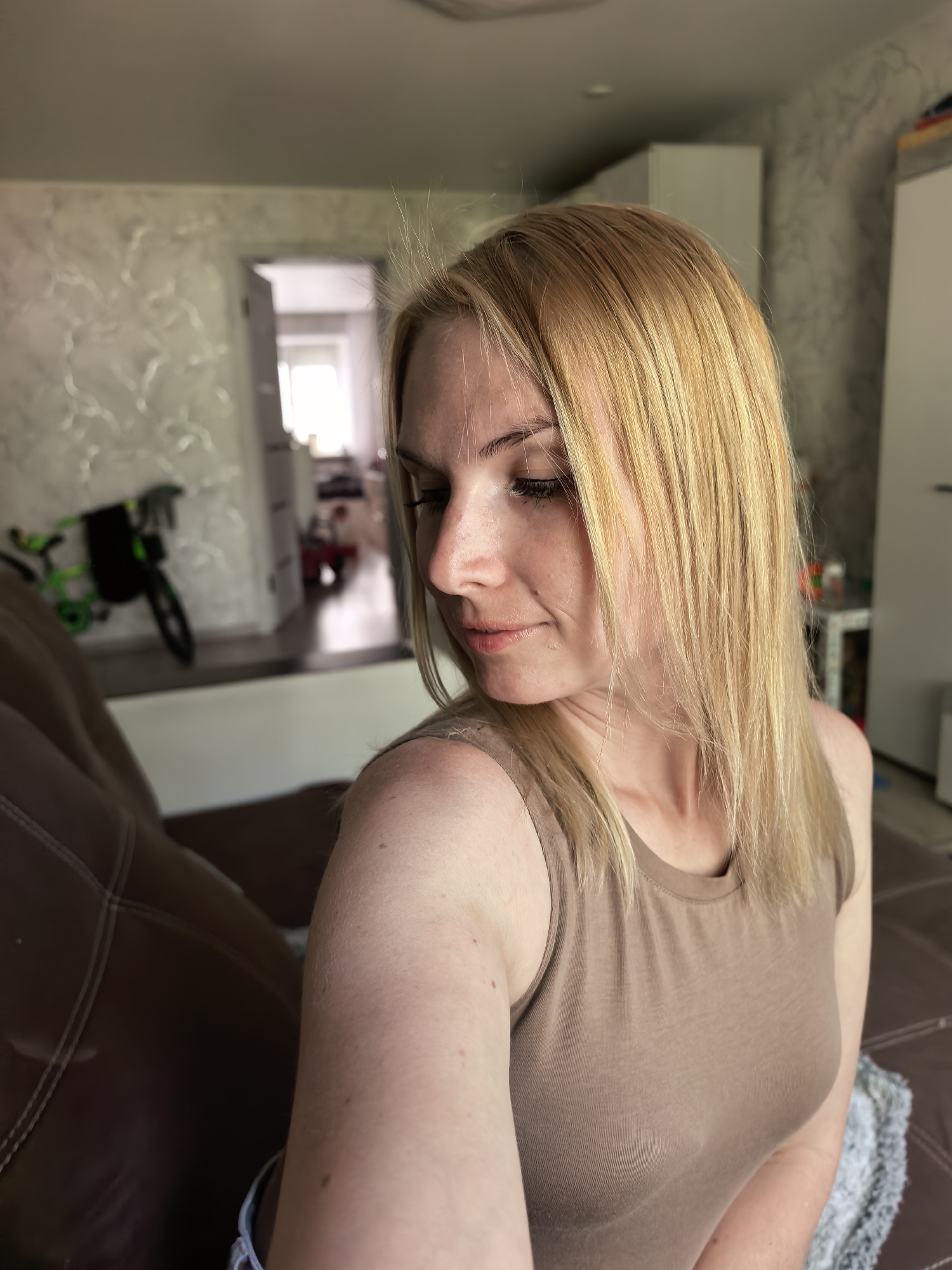 A woman with blonde hair, wearing a brown tank top, sits on a brown leather couch in a room in Sterlitamak, Russia. A child's bicycle is visible in an adjacent room. The walls are decorated with a textured pattern.