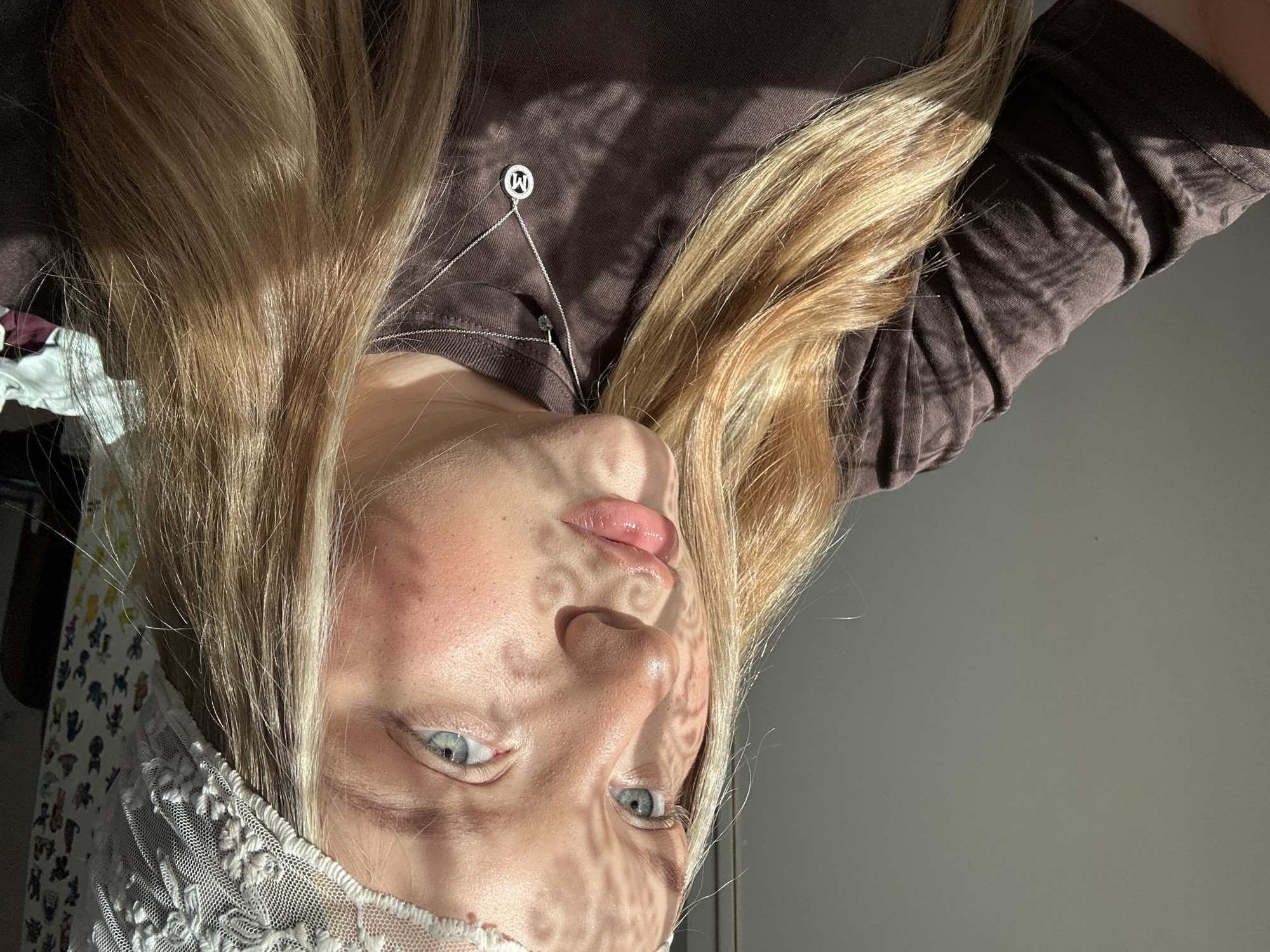 A person with blonde hair and blue eyes is seen from an upside-down perspective. The person is wearing a brown shirt and a white lace headband. A silver necklace with a circular pendant is visible on the person's chest. The person's face is illuminated by sunlight casting a patterned shadow. The background includes a gray wall and a glimpse of other objects. There is no indication this image was taken in Noviy, Russia.