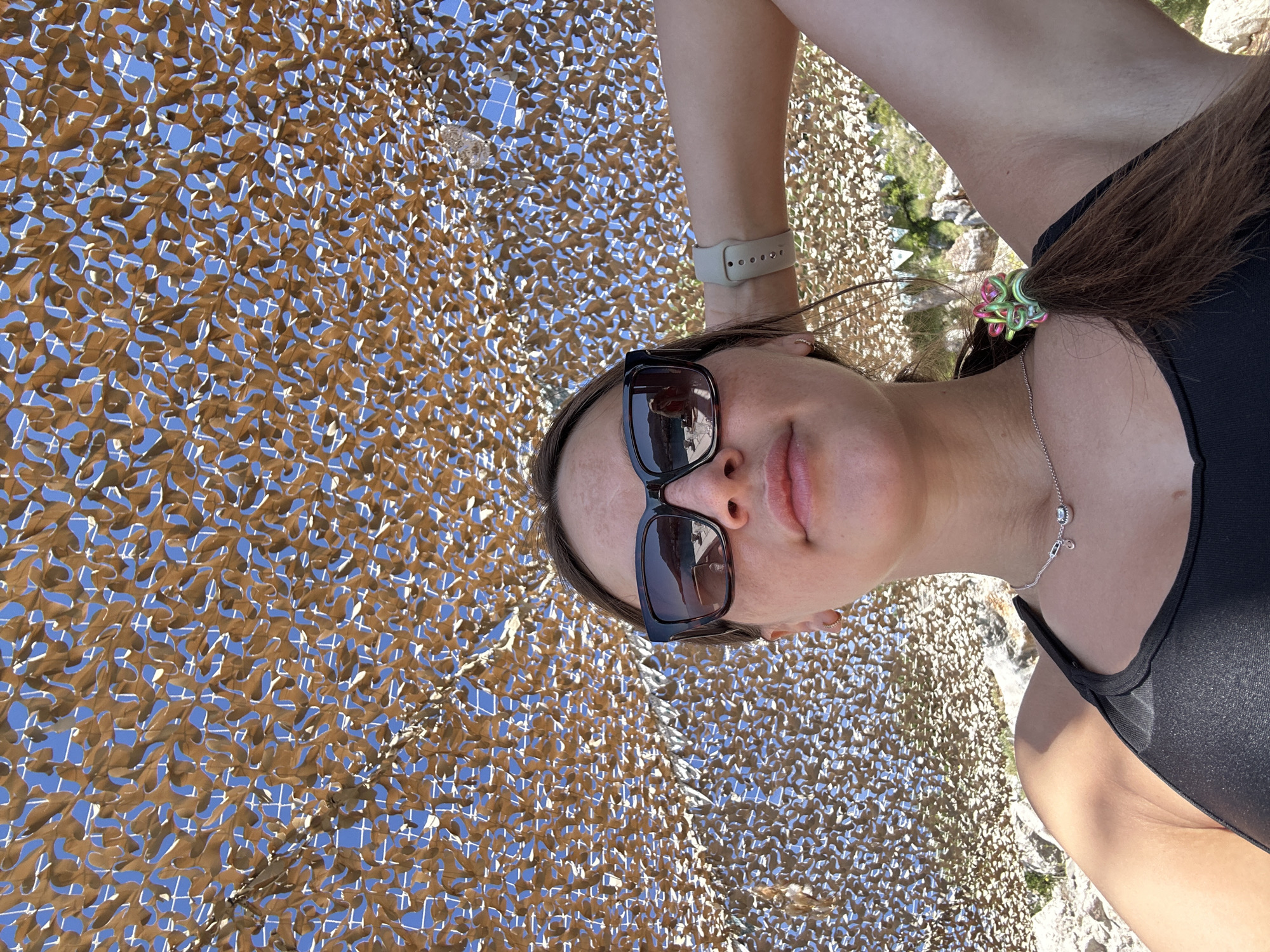 The image shows a young woman in Boreti, Montenegro, taking a selfie from below. She wears sunglasses and a black tank top. Her hair is pulled back with a colorful scrunchie. She is wearing a watch and a necklace with pendant. The background features a brown camouflage netting against a blue sky. Below the netting, partially visible, is a rocky landscape with green vegetation.