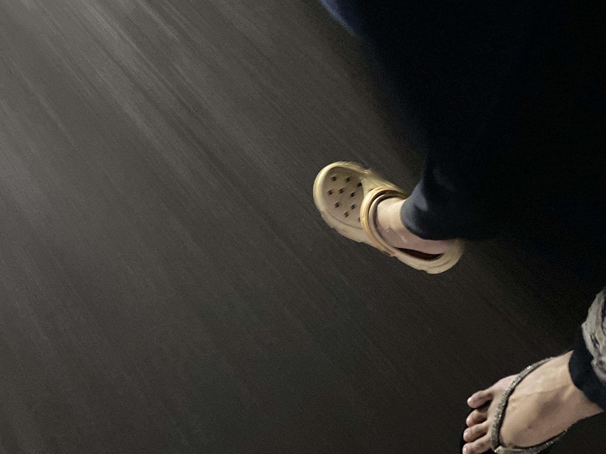 The image shows two feet wearing different footwear against a dark, textured background. The left foot is wearing a sparkly flip-flop and is positioned closer to the foreground. The right foot is wearing a light-colored Croc-style shoe and is positioned further back. The background appears to be a dark, possibly asphalt or concrete surface with visible streaks suggesting motion blur or texture. The lighting is dim, casting a shadow from the right leg. There is no specific indication of the location in Ta', Indonesia.