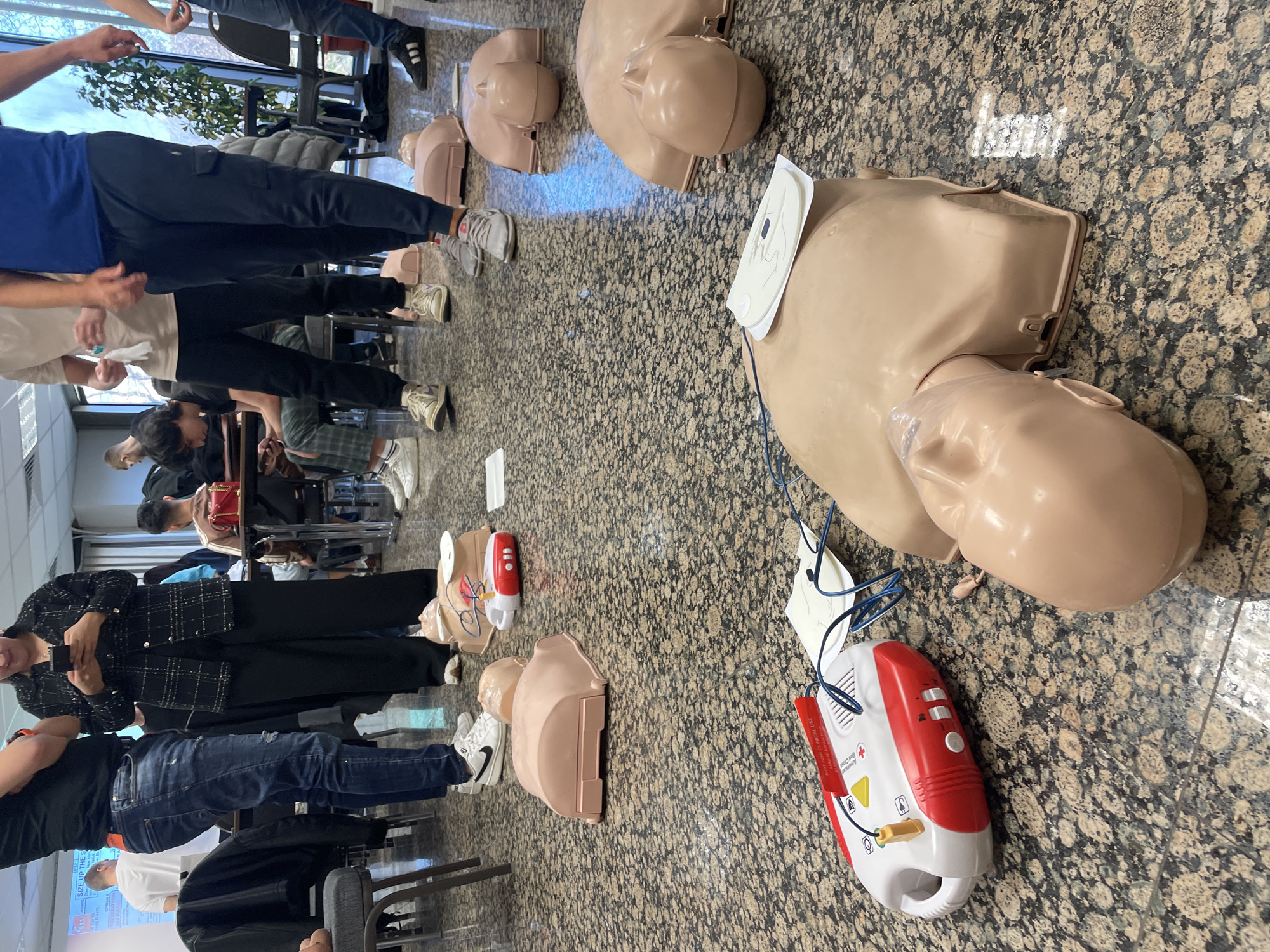 **Scene Description:**
A classroom or training room is visible. The floor is covered in a patterned tile. There are several tan-colored CPR manikins spread across the floor. Some of the manikins have AEDs attached. Several people are standing or sitting around the manikins, seemingly participating in a training session. The location is not identifiable as Sofia, Bulgaria.