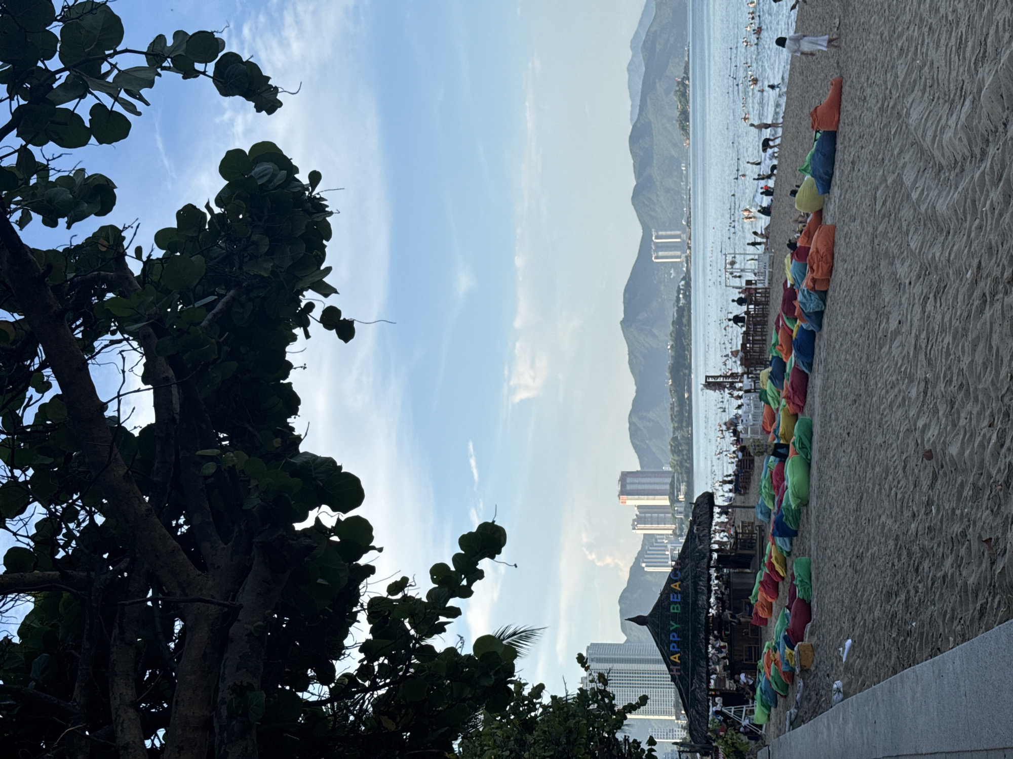 A wide-angle, eye-level shot captures a vibrant beach scene in Nha Trang, Vietnam. In the foreground, a sandy beach is covered with numerous colorful beanbag chairs in shades of orange, green, blue, and red, arranged in rows. A concrete walkway borders the left side of the beach. Towards the left midground, a thatched-roof structure, identified by colorful letters spelling "HAPPY BEACH," functions as a bar or seating area, with people visible under its shade. Beyond the beach, the blue ocean is filled with many bathers. In the background, a cityscape with tall modern buildings and lush green mountains under a partly cloudy blue sky forms the backdrop. A large, leafy tree with dark branches and leaves overhangs the top portion of the image, obscuring some of the sky.