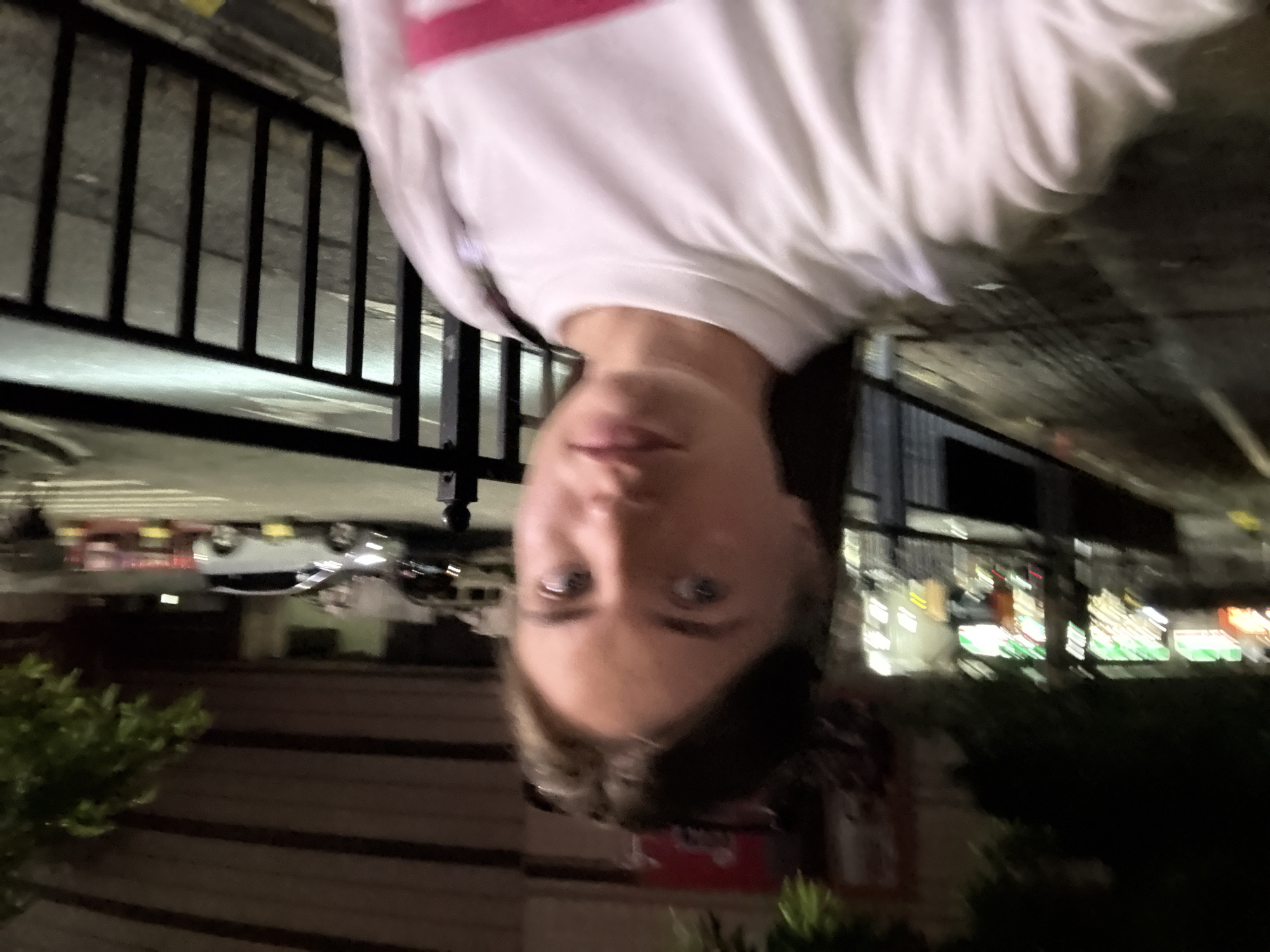 The media file is a close-up, upside-down selfie of a young person in Haizhu District, China. The person is wearing a white long-sleeved shirt with a pink stripe across the chest. Their face is the central focus, with their eyes open and looking towards the camera. The background is blurred, but appears to be an outdoor urban setting at night, with lights from buildings and possibly streetlights visible. There are some dark railings in the foreground, suggesting the person might be on a balcony or near a fence. The overall impression is casual and candid. The image quality is somewhat low, with a soft focus and noticeable grain. No visible text is present in the image.