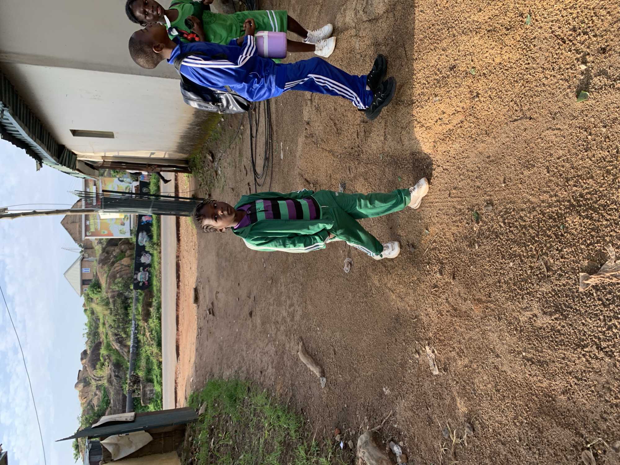 The image depicts a street scene in Jos, Nigeria. In the foreground, a young person in a green tracksuit walks towards the viewer, looking directly at the camera. To their right, two other children are walking away from the viewer; one is wearing a blue tracksuit and carrying a backpack, while the other is wearing a green shirt and carrying a light purple container. The ground is covered in reddish-brown dirt and scattered debris. In the background, a building with a corrugated iron roof is visible, along with a rocky hillside covered in vegetation. The sky is partly cloudy.
