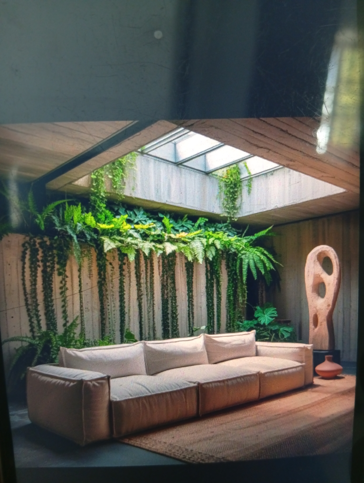 A modern, minimalist interior space is depicted. A large, light beige modular sofa sits on a woven rug, positioned in front of a concrete wall covered in hanging greenery. A skylight provides natural light. To the right is a wooden sculpture and a clay pot. The image suggests an indoor living space with an emphasis on natural elements. There is no evidence to suggest that this image is located in Dutsen Alhaji, Nigeria.
