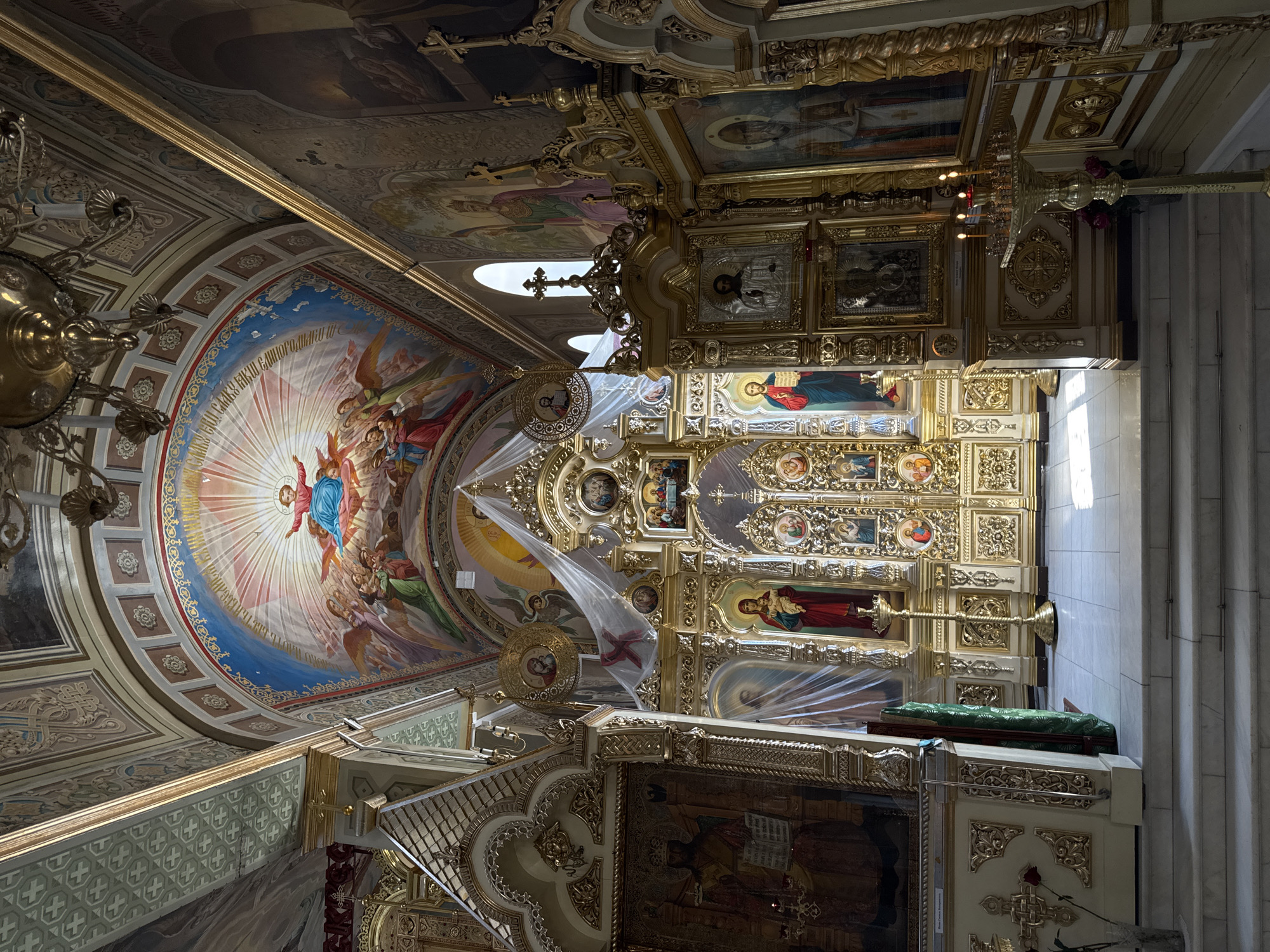 This is a full shot interior view of an Orthodox church. The focal point is the ornate, gold iconostasis, a wall of icons and religious imagery separating the nave from the sanctuary. The iconostasis is intricately carved and gilded, featuring numerous icons of saints and biblical scenes. Above the iconostasis, the ceiling is adorned with a large fresco of the Virgin Mary surrounded by angels. The church interior is richly decorated with religious paintings and gilded elements on the walls and ceiling. Natural light streams in from windows, illuminating parts of the interior and casting shadows. The floor is tiled, and there are marble steps leading up towards the iconostasis. A brass candelabra with lit candles stands in the foreground to the right. The scene conveys a sense of solemnity and grandeur typical of a place of worship.