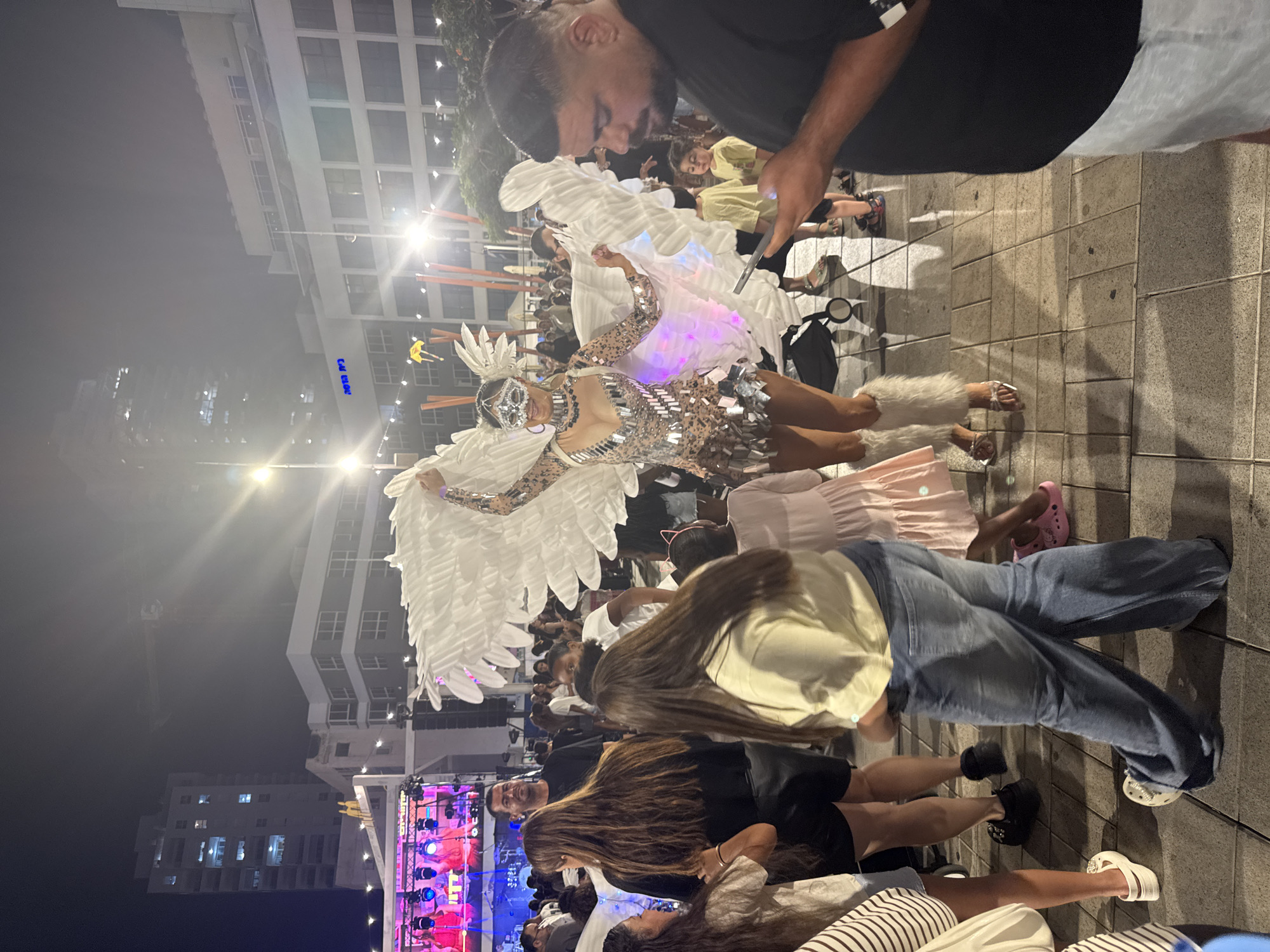 The image captures a nighttime street scene in Netanya, Israel, near the waterfront promenade, characterized by urban architecture and a festive atmosphere. In the foreground, a person dressed in a sparkling, costume adorned with large white wings, resembling an angel or a mythical creature, stands center-frame. This individual wears a jeweled mask and fuzzy leg warmers, and their costume exhibits colorful LED lighting at the bottom.
Surrounding the costumed figure are several people. To the left, a group of women with long hair are visible, one wearing a black top and leather shorts, another in a light-colored sweatshirt and jeans. To the right, a man in a black t-shirt is looking down at his phone, and behind him, two children, a boy in a yellow shirt and another in a blue shirt, are observing the scene. Further back, a crowd of people are scattered throughout the area, suggesting an event or gathering is taking place.
In the background, there are multi-story buildings with illuminated windows, indicating a commercial or residential area. A large stage with colorful lighting and performers can be seen on the far left, suggesting a performance or concert is part of the event. Streetlights and decorative string lights illuminate the scene, casting reflections on the paved ground. The overall environment appears to be an outdoor public space, likely a plaza or street, during a nighttime event.