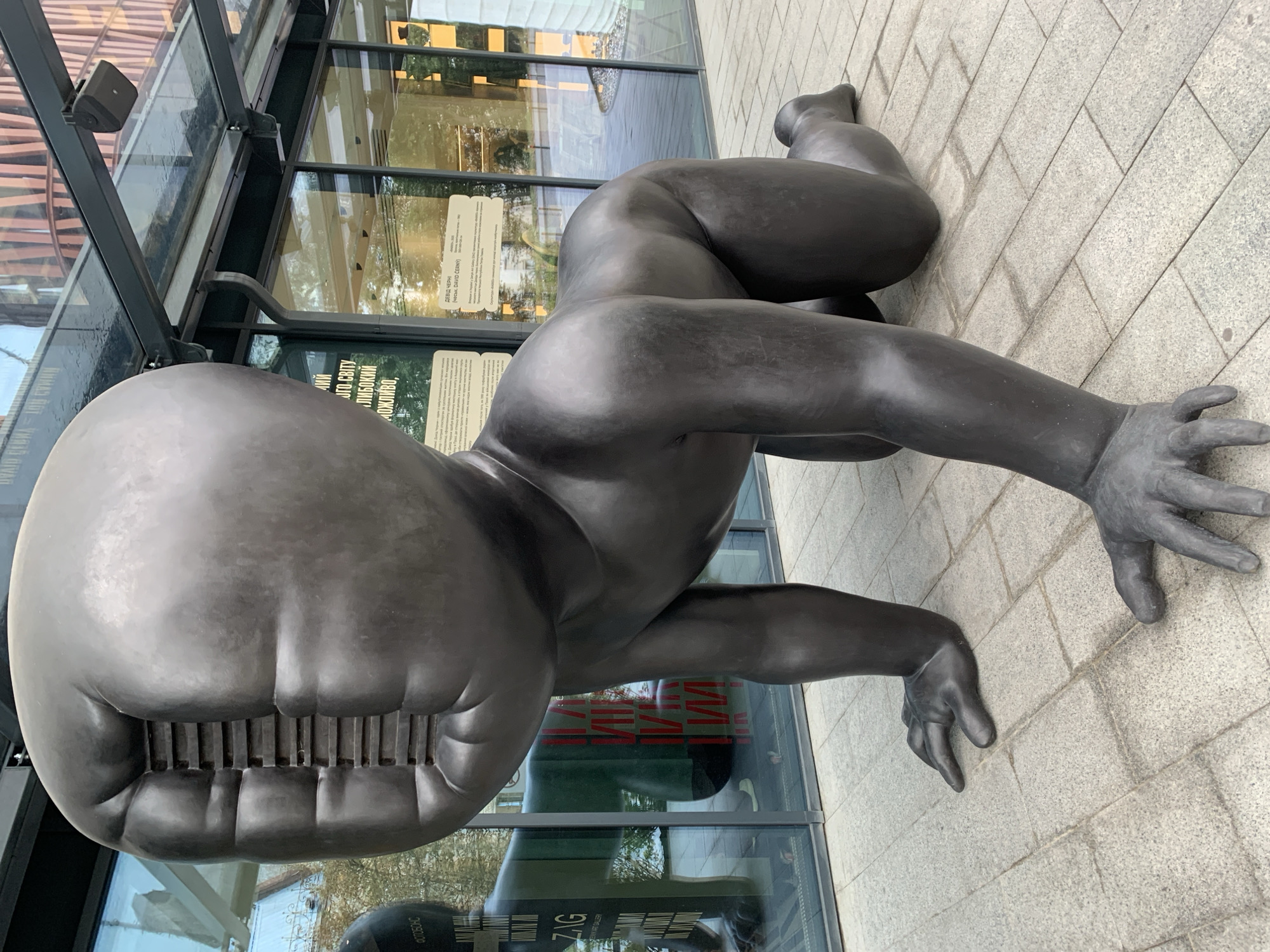 This is a full-shot, eye-level, outdoor photograph of a large, dark bronze sculpture of a crawling baby. The sculpture is located on a paved walkway in front of a modern building with large glass windows and an overhang. The baby sculpture is positioned in the foreground, facing to the left of the frame, with its body angled slightly towards the viewer. Its head is large and bulbous, with a grille-like opening in the face. The baby's limbs are bent as if crawling, and its hands are splayed on the pavement. Reflections of the building and surrounding trees are visible in the glass windows. Two informational plaques are mounted on the exterior of the building behind the sculpture. The overall lighting suggests it is daytime. The sculpture is identified as "The Baby" by David Černý, located outside the Center for Urban History of East Central Europe in Lviv, Ukraine.