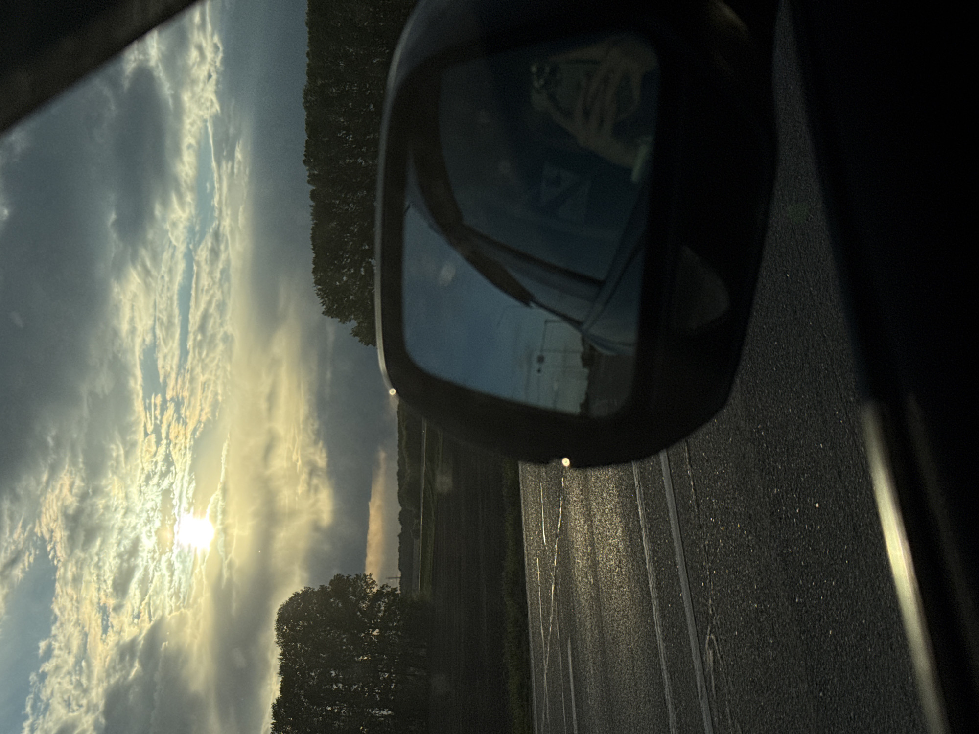The image depicts a view from inside a car, likely while in motion on a road. The focus is on the passenger side-view mirror, which reflects a portion of the sky and possibly buildings or infrastructure. Outside the car window, the sky is visible, showing a bright sun and cloud formations. A portion of the road is visible along with what appears to be vegetation on the side.