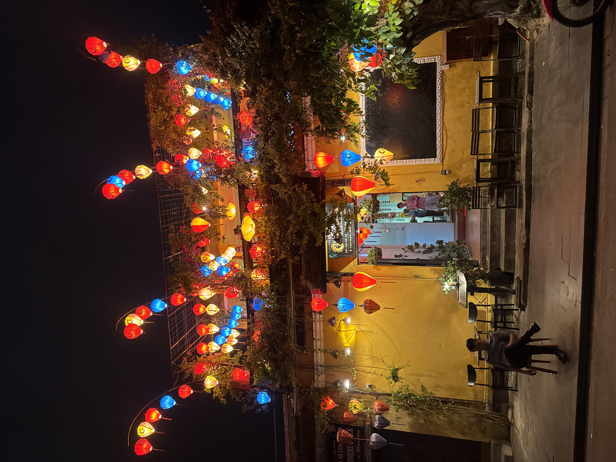 The image shows a scene at night in Hội An Ward, Vietnam. A yellow building is illuminated by colorful lanterns strung across its facade and along the street. The lanterns are red, blue, and yellow, and they cast a warm glow on the surroundings. A person is sitting on a chair outside the building. The person is wearing a gray shirt and dark pants, and they have their legs crossed. Behind them are a few empty chairs and a table. The building has a doorway that leads into a brightly lit interior, where another person is visible. The exterior of the building is decorated with plants and some architectural details. The surrounding area is dark. The text "Cửa Hàng Và Len KHMERE & 2169 93 Bạch Đằng" and "ĐẶC SẢN HỘI AN 75 Bạch Đằng Str, Hội An" is visible on the building.