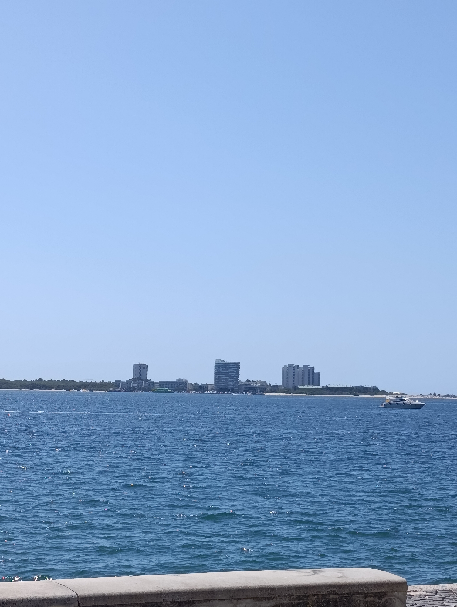 The image shows a panoramic view of the Sado River estuary in Setúbal, Portugal, on a clear, sunny day. The foreground features a concrete barrier, possibly part of a promenade or seawall. The middle ground is dominated by the blue, rippling water of the river, with a white speedboat moving towards the right side of the frame. In the background, a distant coastline is visible, characterized by a line of trees and a cluster of buildings. These include several multi-story structures, some appearing to be modern hotels or residential buildings, and a distinct tall, slender building with visible text on its facade. The sky is a uniform, pale blue, with no clouds. The scene captures a typical seaside urban landscape with a focus on the river and the cityscape.