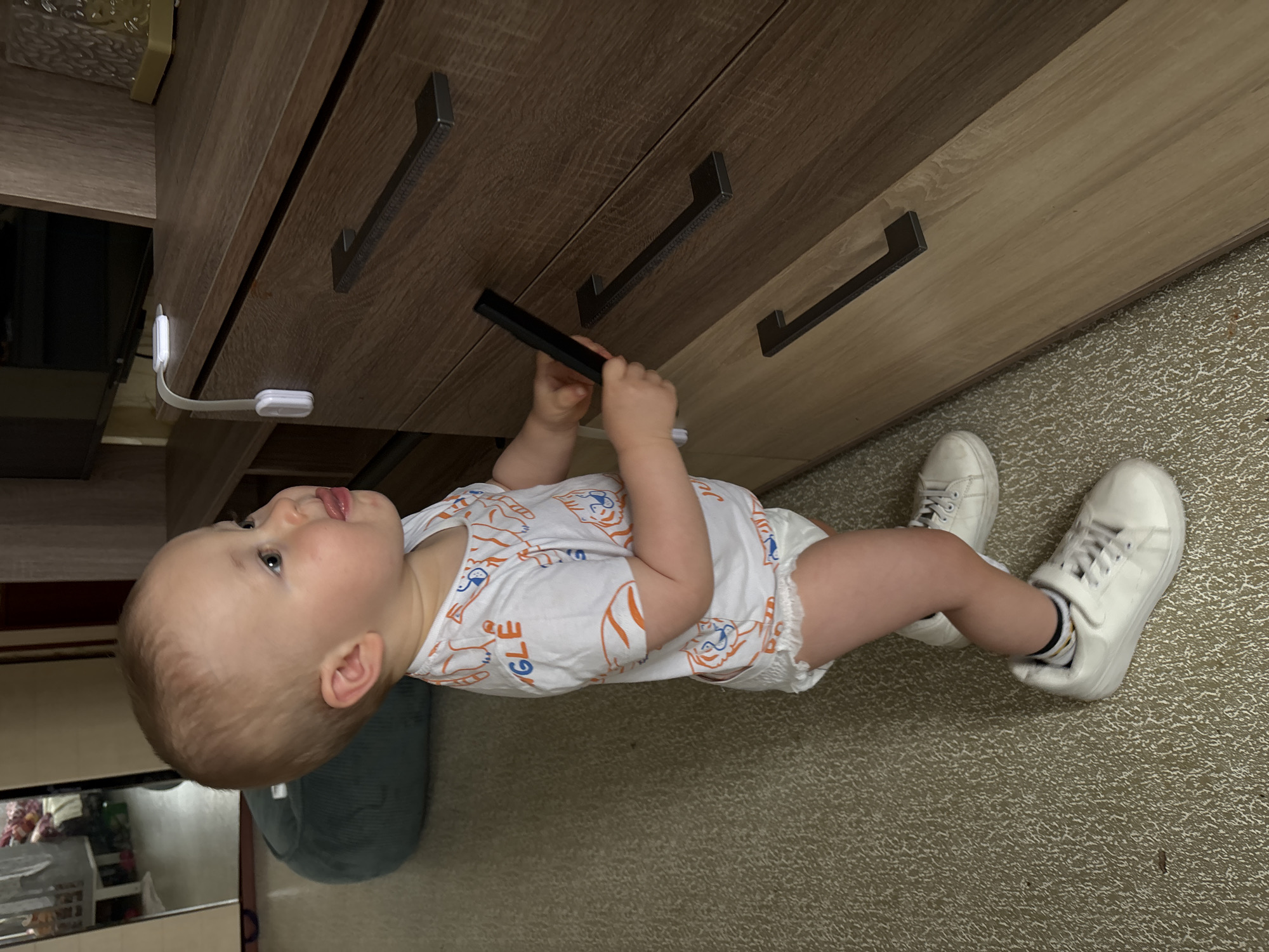 The image depicts a toddler standing on a carpeted floor and reaching towards a wooden dresser. The toddler is wearing a white onesie with an orange tiger print and a white diaper. They are also wearing white sneakers with white laces. The dresser has several drawers with dark handles. A white baby proofing latch is attached to the top drawer. The background shows a room with furniture, including a dark television and a reflective surface which appears to be a mirror or glass door showing part of another room. The scene is indoors. There is no visible indication of the location being Naberezhnye Chelny, Russia.