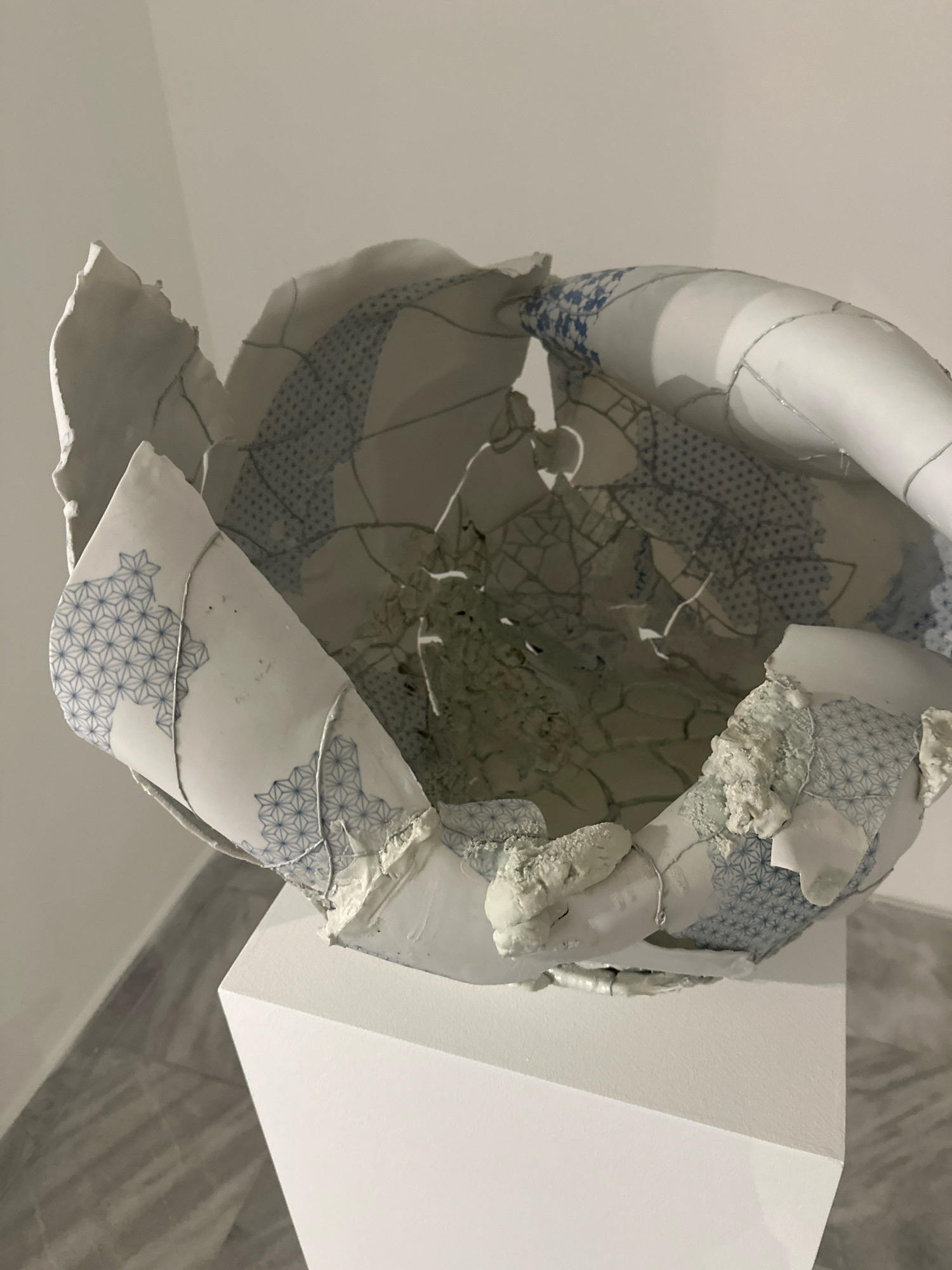 This is a medium-shot, eye-level photograph of a broken ceramic sculpture displayed on a white pedestal. The sculpture, likely a bowl, is shattered into numerous pieces, revealing its fragmented interior. The exterior of the ceramic pieces features a delicate blue and white geometric pattern, reminiscent of traditional porcelain designs. Some fragments are held together by visible white string or wire, suggesting a deliberate attempt at reassembly or artistic reconstruction. The sculpture is positioned centrally in the frame, with its broken edges creating a dynamic, organic shape. The background consists of a plain white wall and a tiled floor, both appearing clean and well-maintained, suggesting an indoor exhibition space. The lighting is soft and diffused, creating subtle shadows that highlight the textures and forms of the sculpture. The overall impression is one of fragility and resilience, with the brokenness of the object serving as a focal point for artistic expression. The context of Belgrade, Serbia, is not visually apparent in the image itself.
