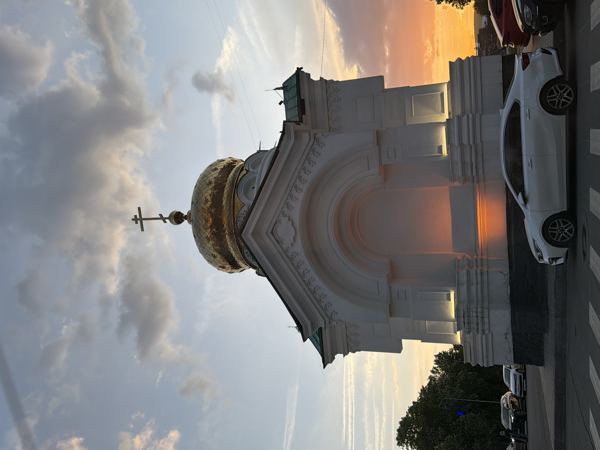 The image depicts a white building with a golden dome topped with a cross, characteristic of Russian Orthodox architecture. The building is situated in Saratov, Russia, as identifiable by the architectural style and surrounding context. The sky is a mix of light blue and orange hues, indicating either sunrise or sunset. Clouds are scattered across the sky.
A white car is parked next to the building. The street is paved with a crosswalk visible. Part of a green tree and other parked cars can be seen in the background, suggesting an urban environment.