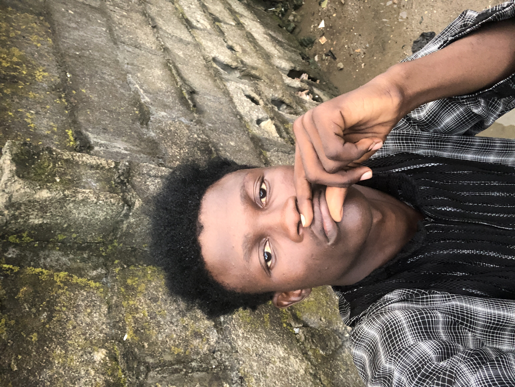 A young Black male, possibly in his late teens or early twenties, is captured in a selfie-style portrait against a rough, grey stone wall. The wall appears weathered and has patches of green moss and lichen, suggesting an outdoor environment. The ground in the background is sandy and uneven, with some scattered debris.
The male has dark, curly hair styled upwards. His skin tone is a deep brown. He is wearing a black and white checkered shirt over a black garment with vertical striped detailing. His right hand is raised, with his fingers gently touching his lips, conveying a contemplative or thoughtful expression. His gaze is directed towards the camera. The lighting suggests it is daytime, likely overcast or in shade, as there are no harsh shadows. The overall impression is a casual, unposed moment in an urban or semi-urban setting, possibly Ado, Nigeria.
