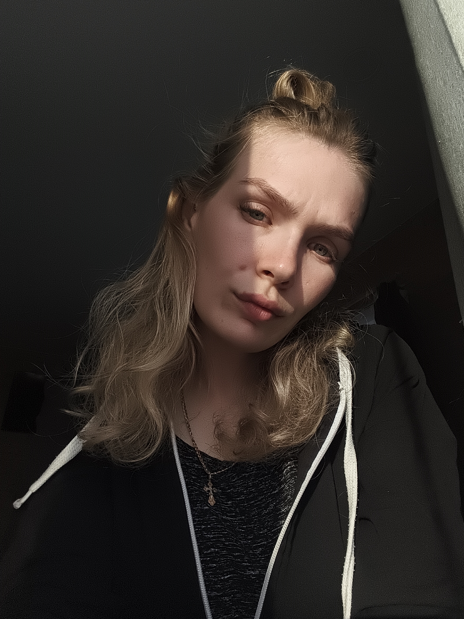 A fair-skinned young woman with blonde hair pulled back into a partial bun, is captured in a medium close-up shot. Her face is partially illuminated by sunlight, while the rest of the image is in shadow. She wears a black hoodie with white drawstrings over a dark gray patterned top. A gold necklace with a pendant is visible. The background is a mix of dark and bright areas, appearing to be an interior space. There is no specific evidence to identify the location as Русский Вожой, Russia.
