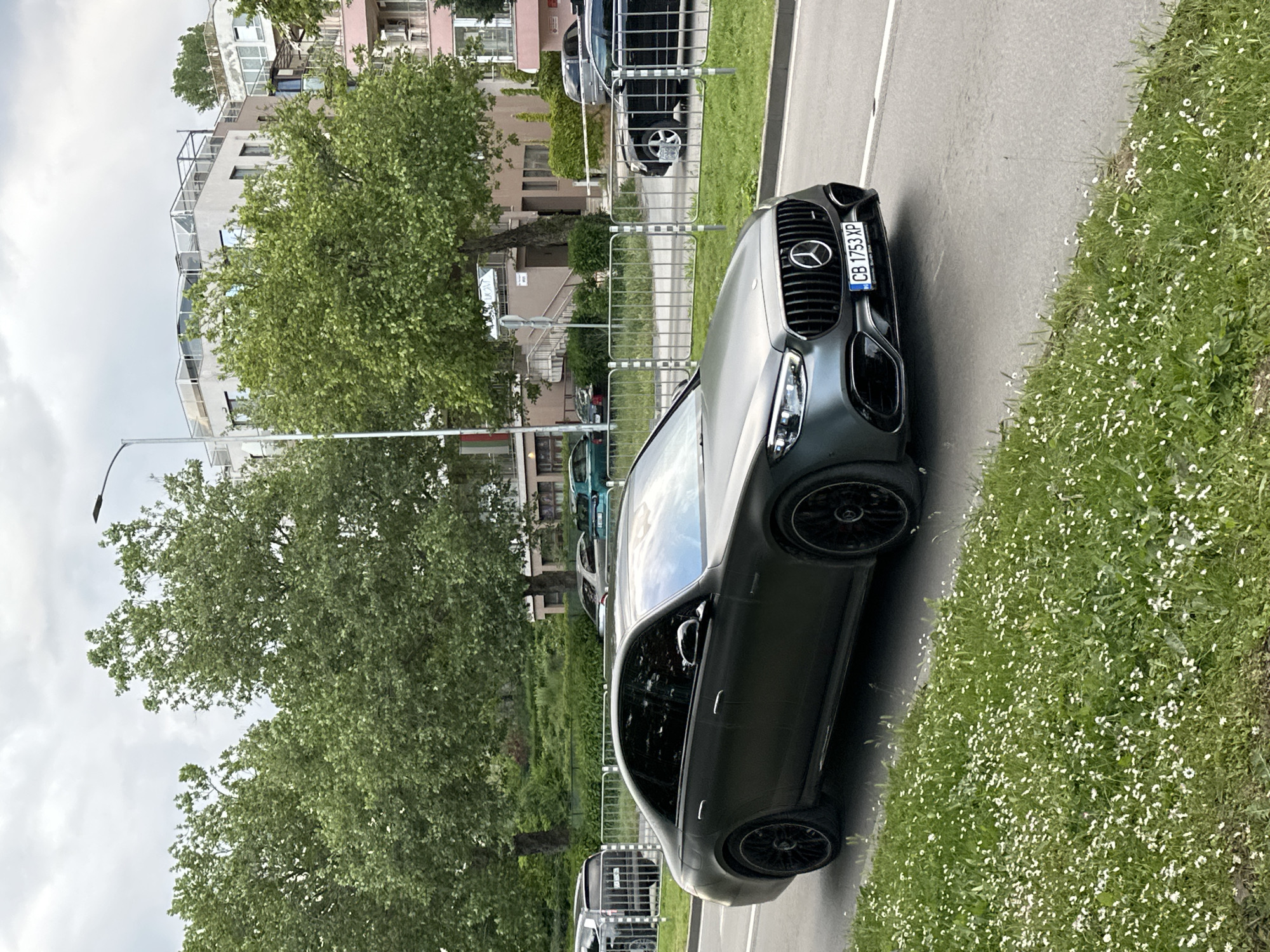 The image depicts a charcoal gray Mercedes-Benz SUV parked on a paved surface next to a grassy area filled with small white flowers. The car's license plate, visible on the front grille, reads "CB 1753 XP." In the background, there is a row of apartment buildings behind a metal fence and several trees, including a large tree with dense green foliage obscuring some of the buildings. The location appears to be a street in an urban area, likely in Varna, Bulgaria, based on the license plate. There are also other cars parked in the background. The weather appears to be overcast.