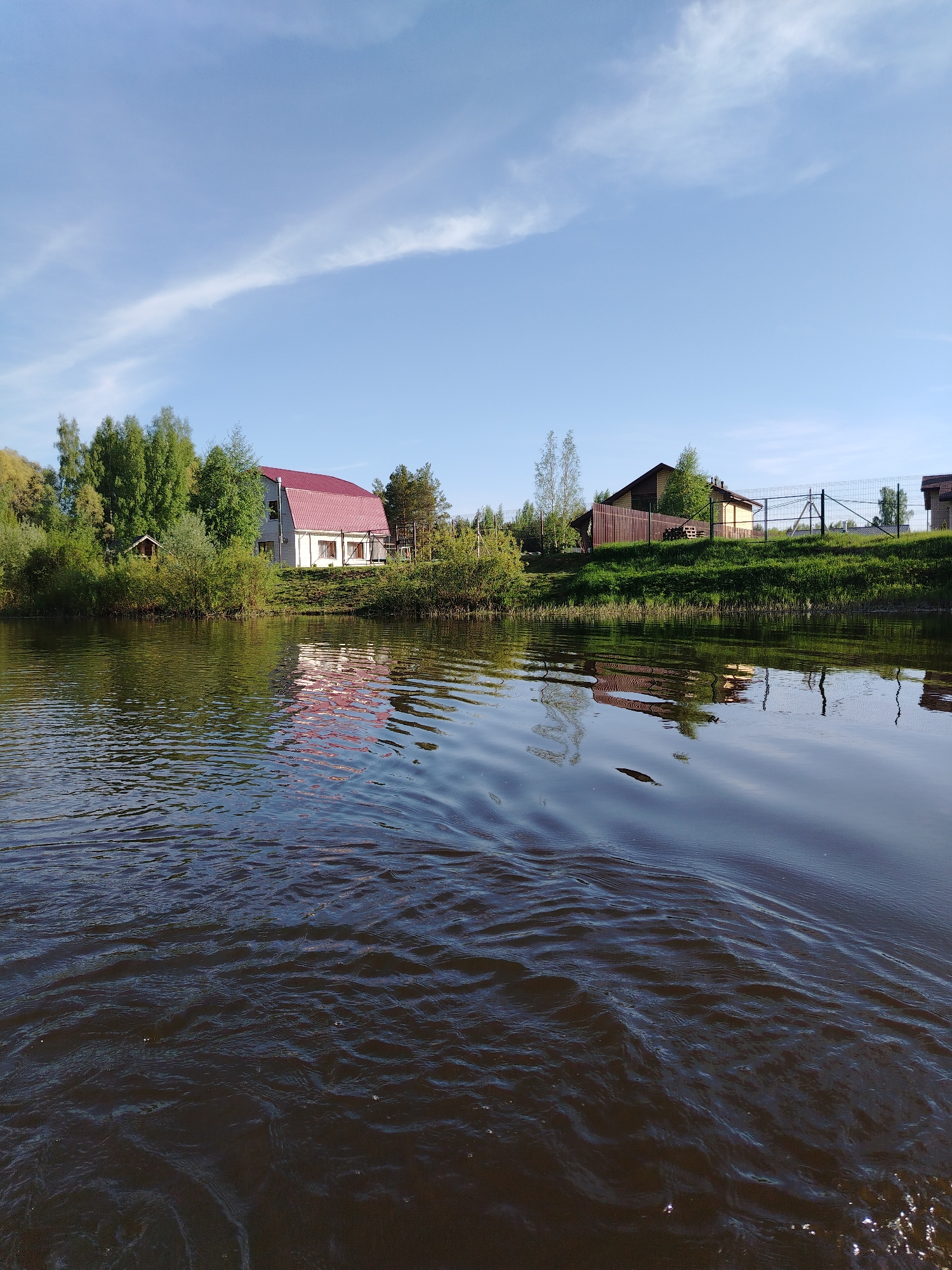 The image depicts a body of water, likely a lake or wide river, in Stroitel', Russia. The water is dark and rippled, with reflections of the shoreline visible. On the shoreline, there is a mix of vegetation, including trees and bushes. Several buildings are visible along the shoreline, including a white building with a red roof and a brown building with a fence. The sky is blue with some wispy clouds. The overall scene suggests a rural or suburban setting in Stroitel'.