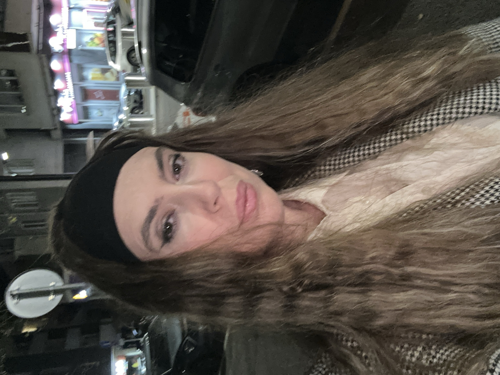 A woman with long brown hair and a black headband is shown in a selfie taken at night in Tbilisi, Georgia. She is wearing a black and white patterned coat and a light-colored shirt or scarf underneath. The background shows city lights, including a brightly lit storefront, and a dark-colored car. A street sign is visible in the lower left corner.