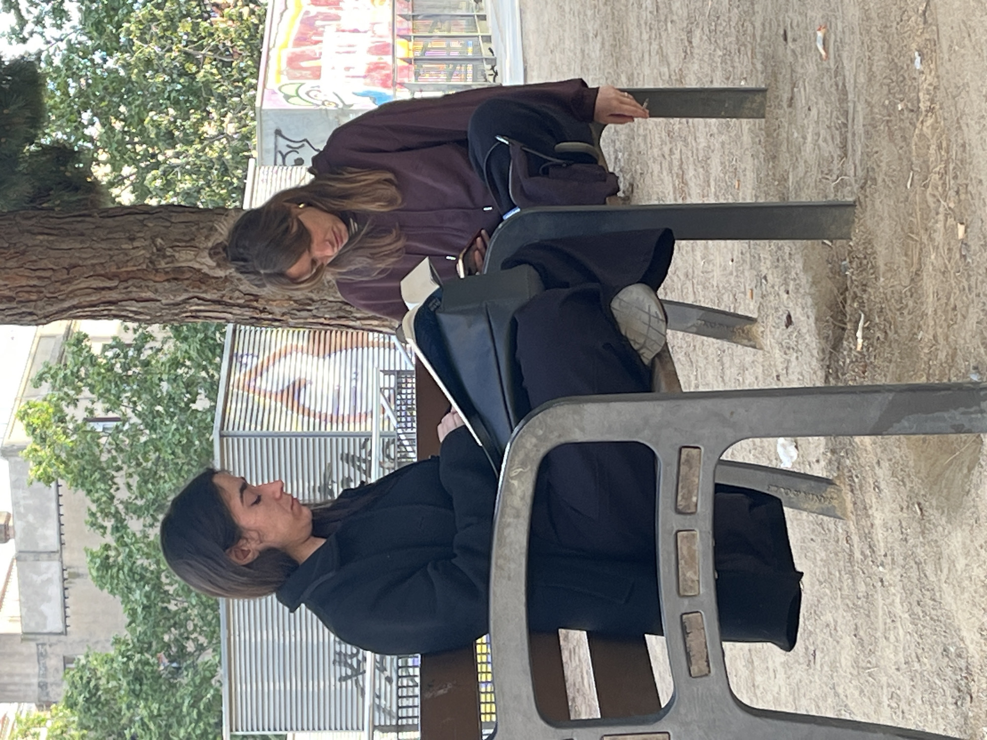 The image, which is originally rotated 90 degrees clockwise, captures two young women seated on outdoor benches in what appears to be a public park or plaza, likely in Barcelona, Spain.
In the foreground, a woman with dark hair, wearing a black jacket or hoodie, is seen seated on a grey metal bench. Her head is tilted downwards, and her eyes are mostly closed, suggesting a moment of quiet contemplation or focus on something in front of her. Her hands rest on her lap.
Further back, on an adjacent bench, another woman with lighter brown hair is visible. She wears a burgundy-colored hoodie or jacket and is also looking down, presumably at a personal device or item held in her hands. A thin, light-colored object, appearing to be a cigarette, is held between the fingers of her right hand. A dark-colored backpack or bag rests on the bench between the two individuals.
The setting includes a large tree trunk with textured bark on the left side of the frame, extending upwards into green foliage. The ground beneath the benches is composed of light-colored sand or gravel. In the background, a vibrant graffiti-covered wall adds an urban artistic element, displaying bright colors such as red, yellow, blue, and green, along with abstract shapes and letter-like forms. Beyond the wall and trees, glimpses of building structures suggest an urban environment.
The scene takes place during daylight hours, indicating mild weather. The women appear relaxed and absorbed in their individual activities. Faintly visible on the side frame of the foreground bench is the text 'SANTANA', possibly accompanied by a year such as '2006' or '2000'.
