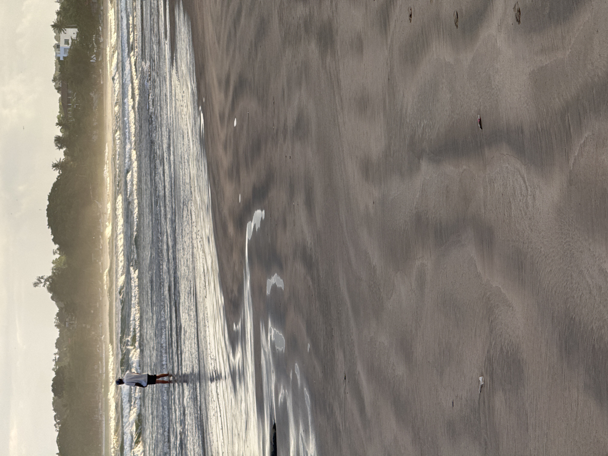 A person stands in shallow water on a beach, facing away from the camera. The person is wearing a light-colored short-sleeved shirt and dark shorts, with a dark baseball cap. The waves are moderate, with whitecaps, and extend to the horizon. In the background, there is a tree-lined coastline with some buildings visible on a hill. The foreground shows wet sand with ripple patterns left by receding water. The overall lighting suggests it is either early morning or late afternoon. The scene is the Weligama beach in Weligama, Sri Lanka.