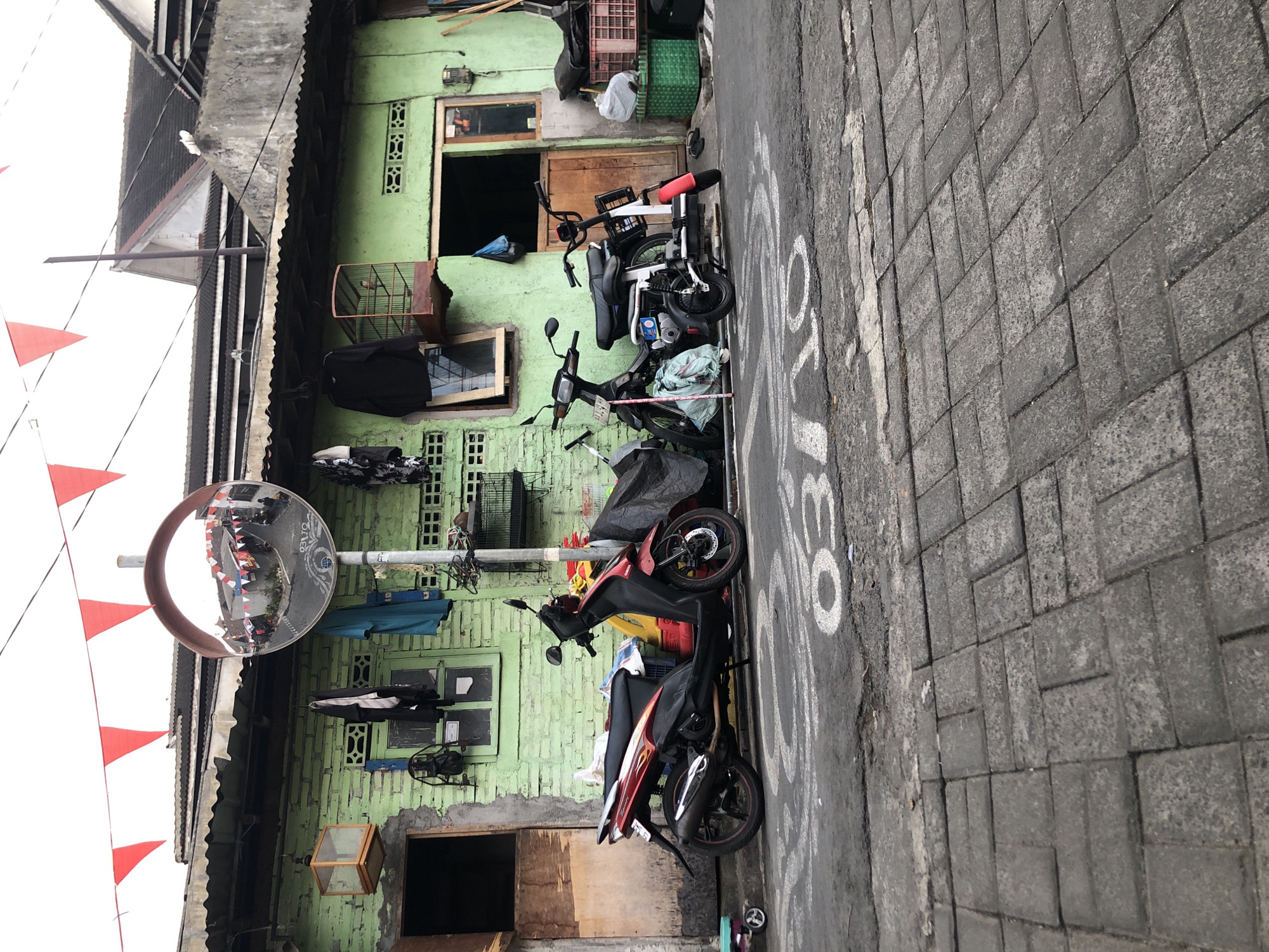 The image depicts a street scene in Magelang, Indonesia, characterized by a simple, lime-green brick building with a corrugated metal roof. Red and white triangular flags are strung across the top of the frame, suggesting a festive occasion. In front of the building, two motorcycles are parked: a red and silver scooter on the left and a smaller, white electric motorcycle on the right. A convex mirror on a pole reflects the street and buildings opposite, showing red and white flags similar to those in the foreground. Several items are hung from the building, including clothes and bird cages, and there are open doorways and windows. The street is paved with dark grey cobblestones in the foreground and asphalt further back, marked with white painted numbers "03" and a circular symbol. The overall impression is of a well-worn but lively urban environment.