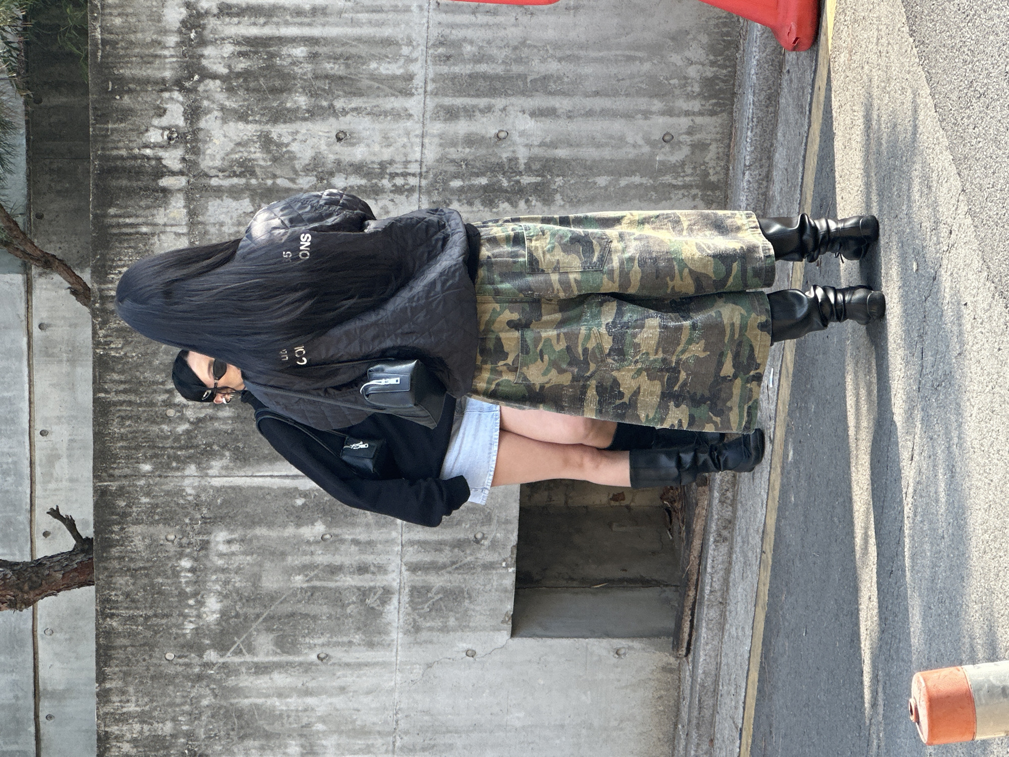 The image depicts two individuals standing outdoors in an urban setting identified as Seoul, South Korea. The scene is illuminated by natural light, casting shadows on the asphalt surface in the foreground.
In the foreground, a person with long, straight black hair is viewed from the back, wearing a black quilted bomber jacket with white text partially visible on the back ("ONS"), oversized camouflage-print wide-leg pants, and black leather boots. This individual also wears a small black shoulder bag diagonally across their back and is positioned on the edge of an asphalt road, facing towards a concrete wall in the background.
To the left and partially obscured by the first individual, a second person is visible. This individual has dark hair pulled back, is wearing dark sunglasses, a black long-sleeved top, a light-wash denim mini-skirt, and black tall boots. They also carry a small black shoulder bag. Their head is turned slightly to the left, away from the immediate foreground.
The background features a textured concrete wall with visible circular formwork holes and dark stains. Above the wall, the trunk and branches of a bare tree are visible. A rectangular recessed opening is present at the base of the concrete wall on the left. The ground consists of asphalt with a yellow painted line, transitioning to a concrete curb where the individuals are standing. In the bottom left foreground, a gray and orange traffic bollard is present on the asphalt. A red plastic barrier or cone is partially visible on the right edge of the frame, near the concrete wall.