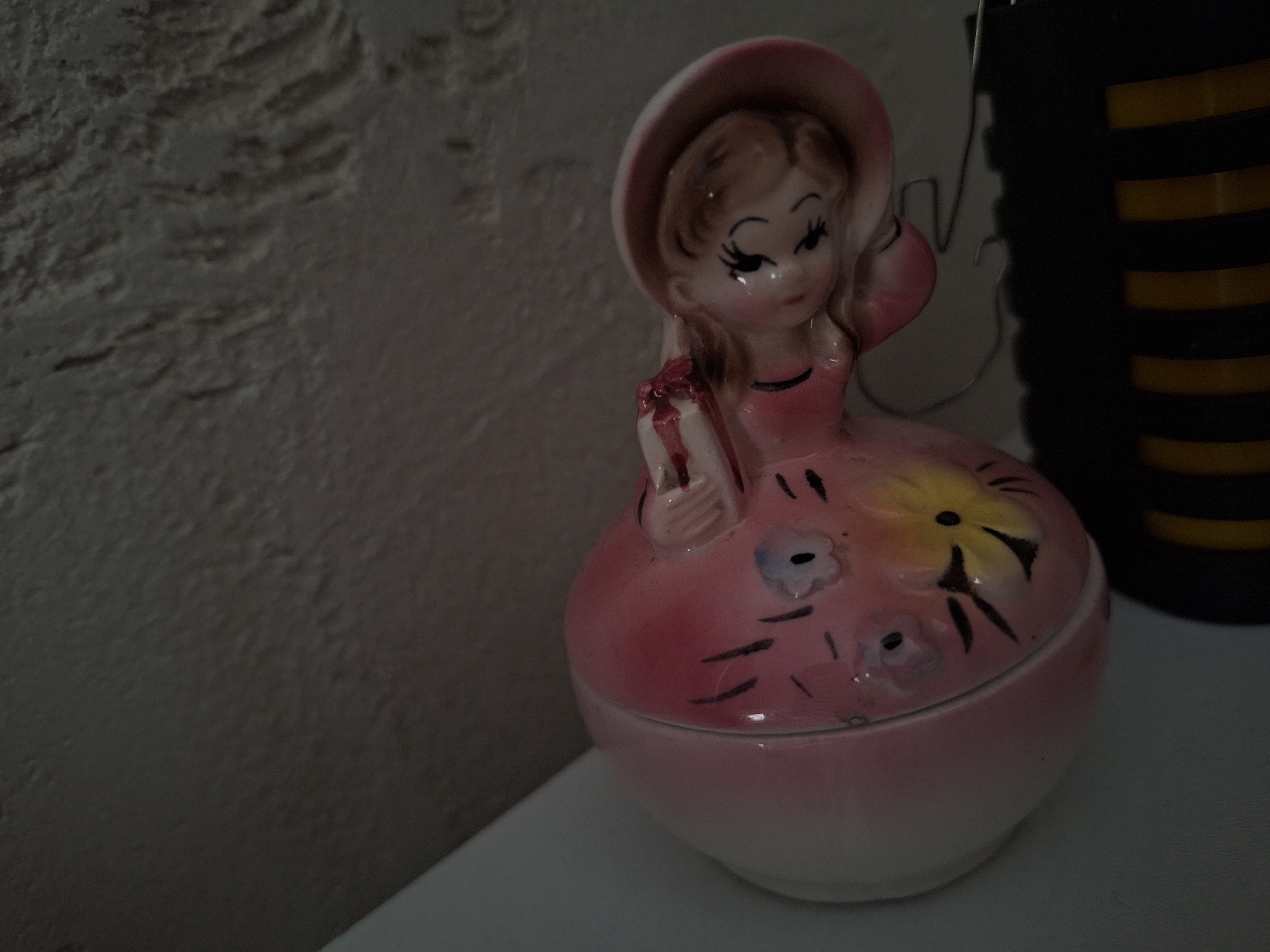 A vintage porcelain figurine of a girl wearing a hat and dress with floral decorations is on a white surface. The girl holds a small, rectangular object. A textured wall is in the background to the left, and a black and yellow striped object stands to the right of the figurine. There is no evidence to suggest the location is in Panagyurishte, Bulgaria.